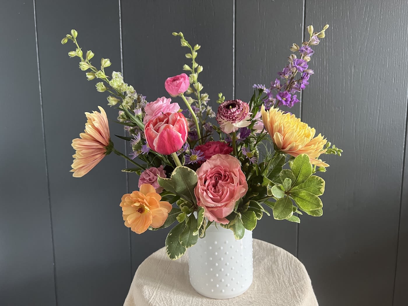 Mixed bouquet of pink, peach, yellow, and purple flowers in a white vase