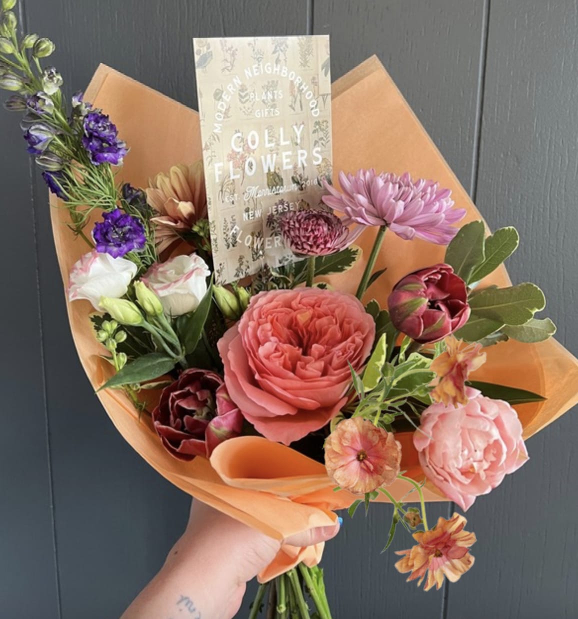 Hand-tied bouquet of pink, purple, and peach flowers wrapped in orange paper