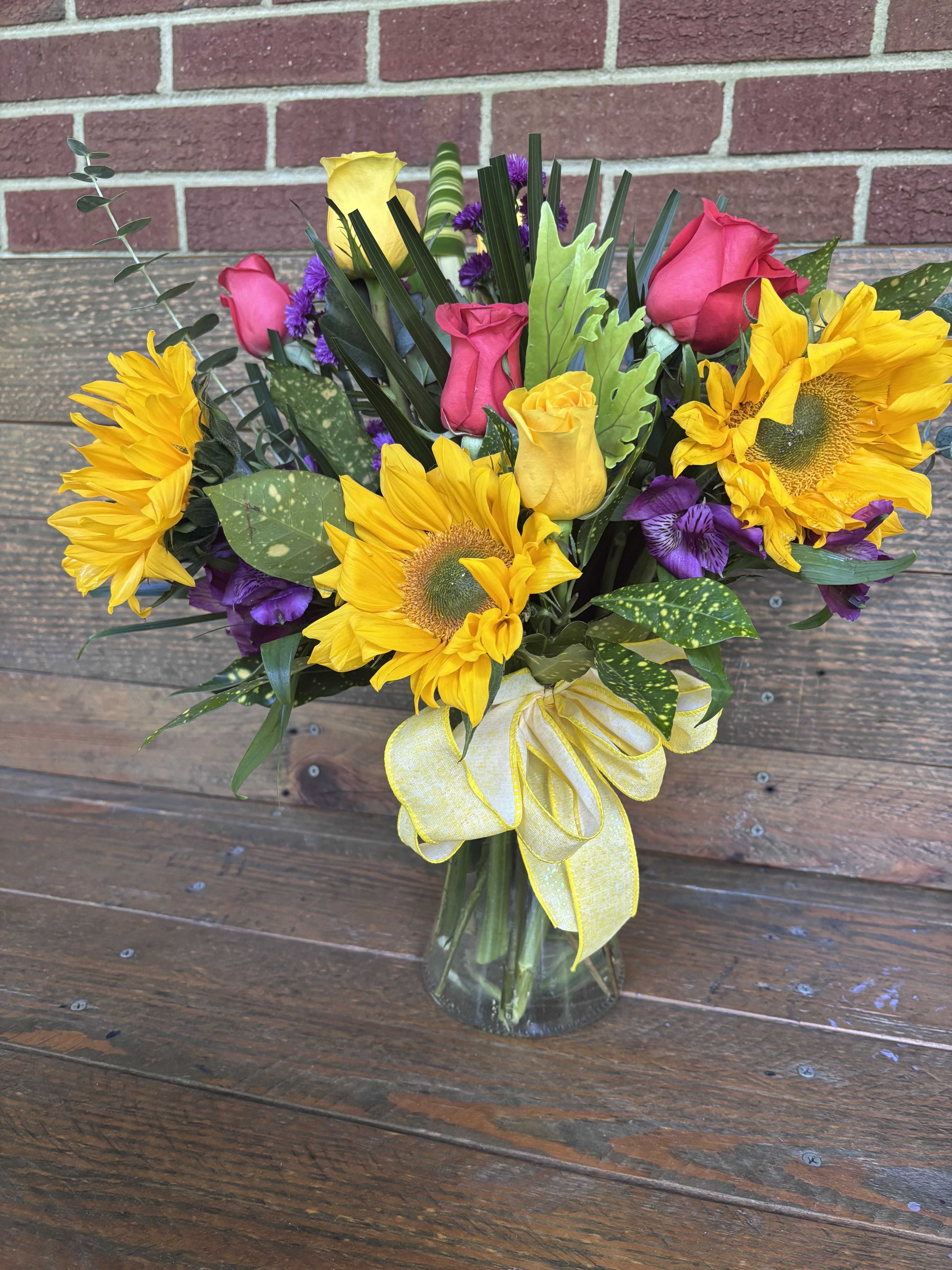 Solana - Sunflowers take center stage in this cheerful flower arrangement, paired with pink and yellow roses for a warm, uplifting look perfect for a birthday bouquet, casual celebration, or a bright "thank you" gift. The Solana arrangement is designed to make the person you're thinking of feel noticed and celebrated-the bold sunflowers create an immediate smile while the softer rose tones add affection and charm.  This full, layered design mixes vivid yellow sunflowers, pink and pale yellow roses, and small purple filler blooms set in varied green foliage for texture and depth. The flowers arrive gathered in a clear glass vase and are finished with a large yellow ribbon tied at the base, giving the piece a polished, gift-ready appearance with a slightly rustic, hand-arranged feel. The combination of sunny yellows, lively pinks, and subtle purple accents creates a vibrant contrast that reads as joyful and upbeat.  Perfect for everyday gifting, hostess visits, or brightening someone's desk or kitchen table, Solana is an easy way to lift a mood and celebrate small moments. Arrives ready to display in the included clear glass vase; tuck stems in fresh water and trim as needed to prolong bloom life.