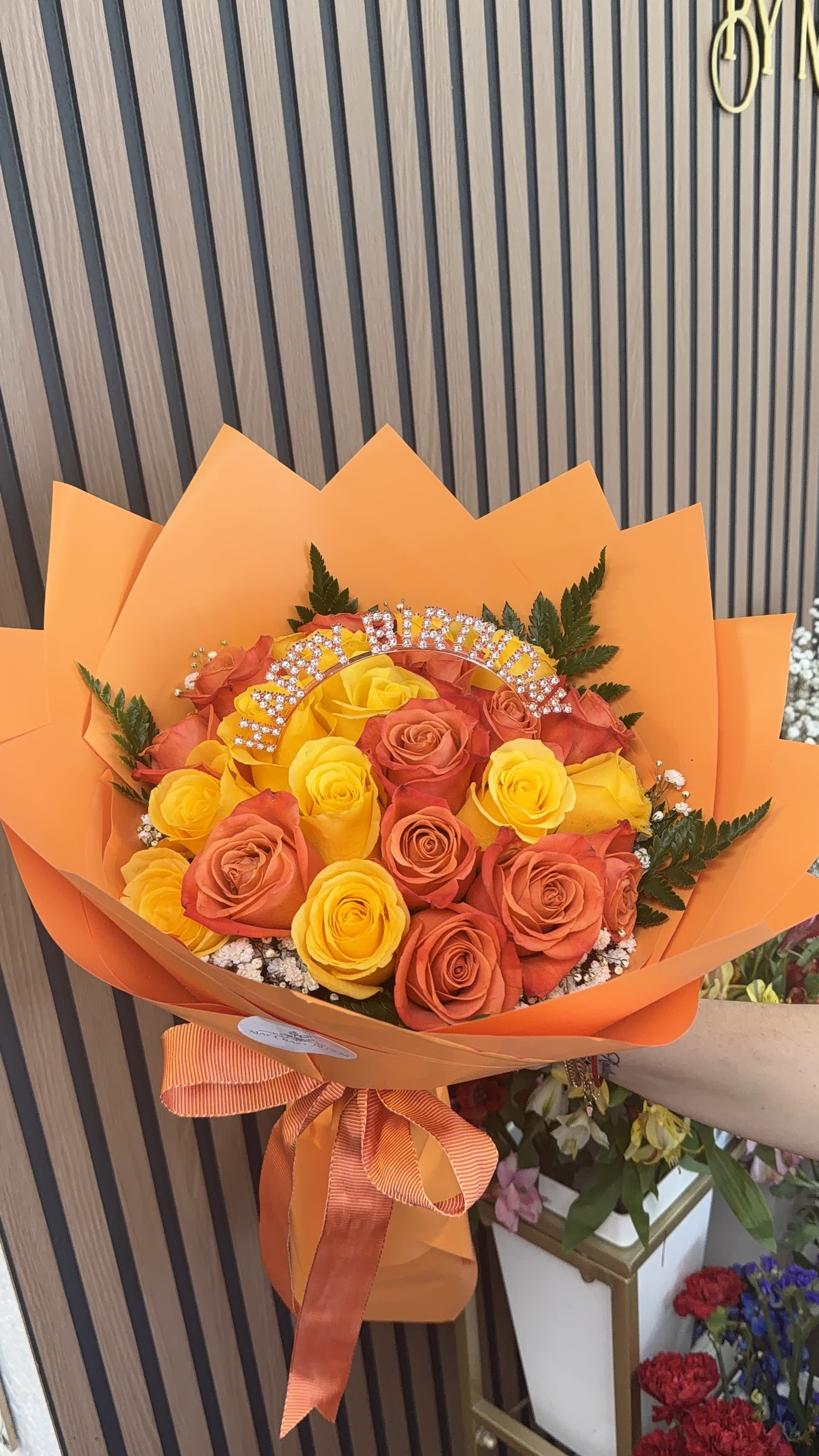 24 Orange wrap of roses  - Sunshine-bright and celebratory, this orange wrap of roses is a joyful birthday bouquet that makes the recipient feel truly celebrated. Tightly packed yellow and orange roses form a rounded, hand-tied spray that reads as cheerful and vivid-perfect for birthdays, congratulations, or any upbeat "thinking of you" moment when you want someone to smile.  The arrangement combines vivid orange, yellow and hints of red blooms with dark green foliage and tiny white filler flowers for contrast. Bright orange wrapping paper and a coordinating striped ribbon amplify the warm palette, while a decorative rhinestone birthday topper arcs above the blooms for an extra touch of sparkle. Available in three sizes-12, 24, and 50 roses-this bouquet arrives ready to display and brighten any room.  Designed for gifting, she'll feel noticed and celebrated the moment the bouquet arrives; the rhinestone topper makes it especially fitting for milestone birthdays or festive table accents. To keep the roses looking fresh, place the stems in clean water and trim ends every few days; select your preferred rose count at checkout to match the occasion and budget.