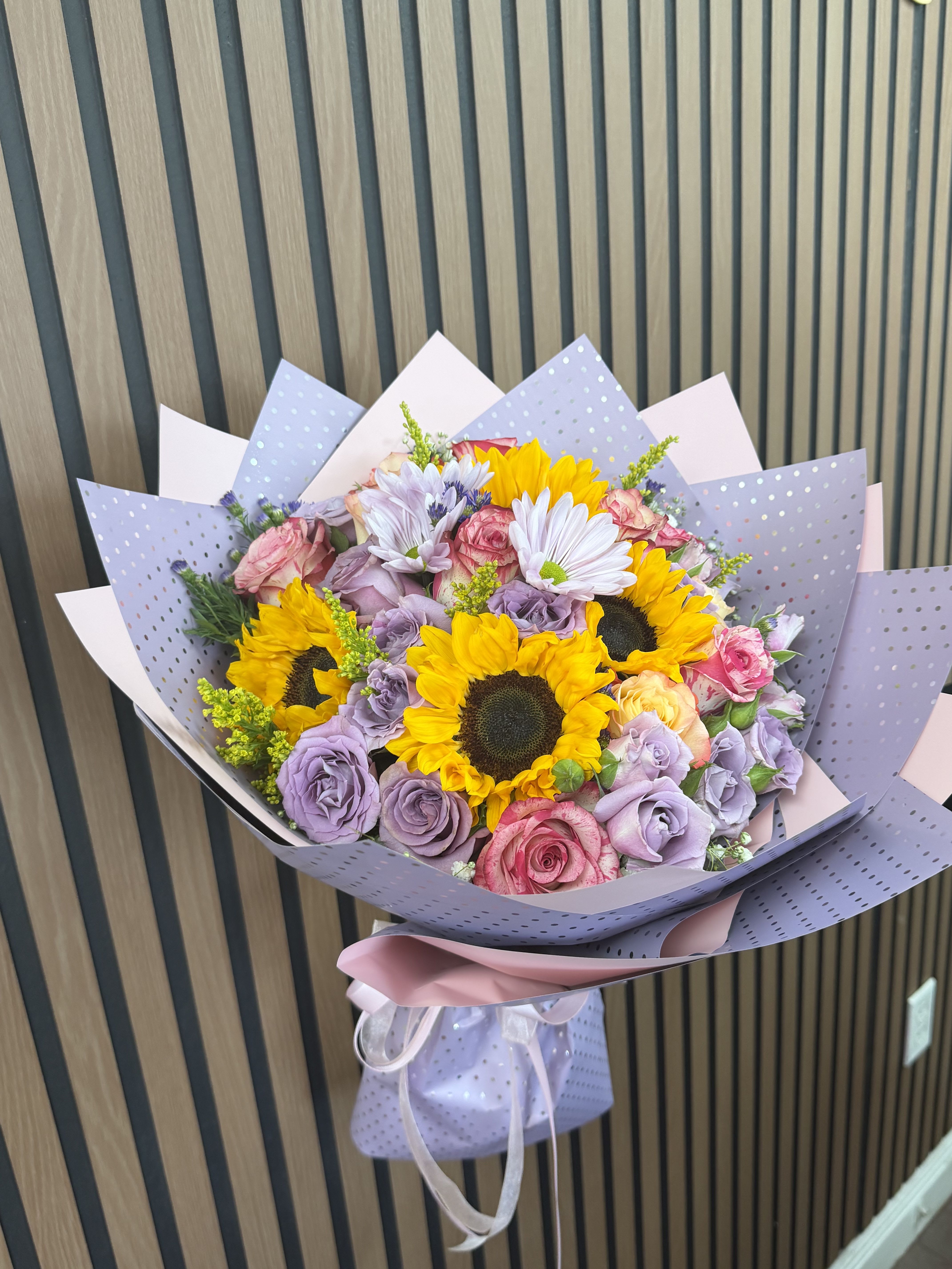 Wonder - Bright sunflowers steal the show in this cheerful flower arrangement, balanced by lavender and pink roses and crisp white daisy-like blooms-perfect as a birthday bouquet, congratulations gift, or a cheerful everyday surprise. The mix of warm sun-kissed yellow with cool lavender and soft pink creates an uplifting, celebratory impression that will make the person you're thinking of feel truly seen and celebrated.  This full, rounded arrangement layers large sunflower heads with spray roses and daisies for texture and gentle movement. Pastel lavender and pink decorative wrapping paper and a neat ribbon finish the presentation, giving it a soft, gift-ready look that's easy to place on a table or desk. Order it to brighten someone's day; the arrangement arrives ready to display and enjoy.