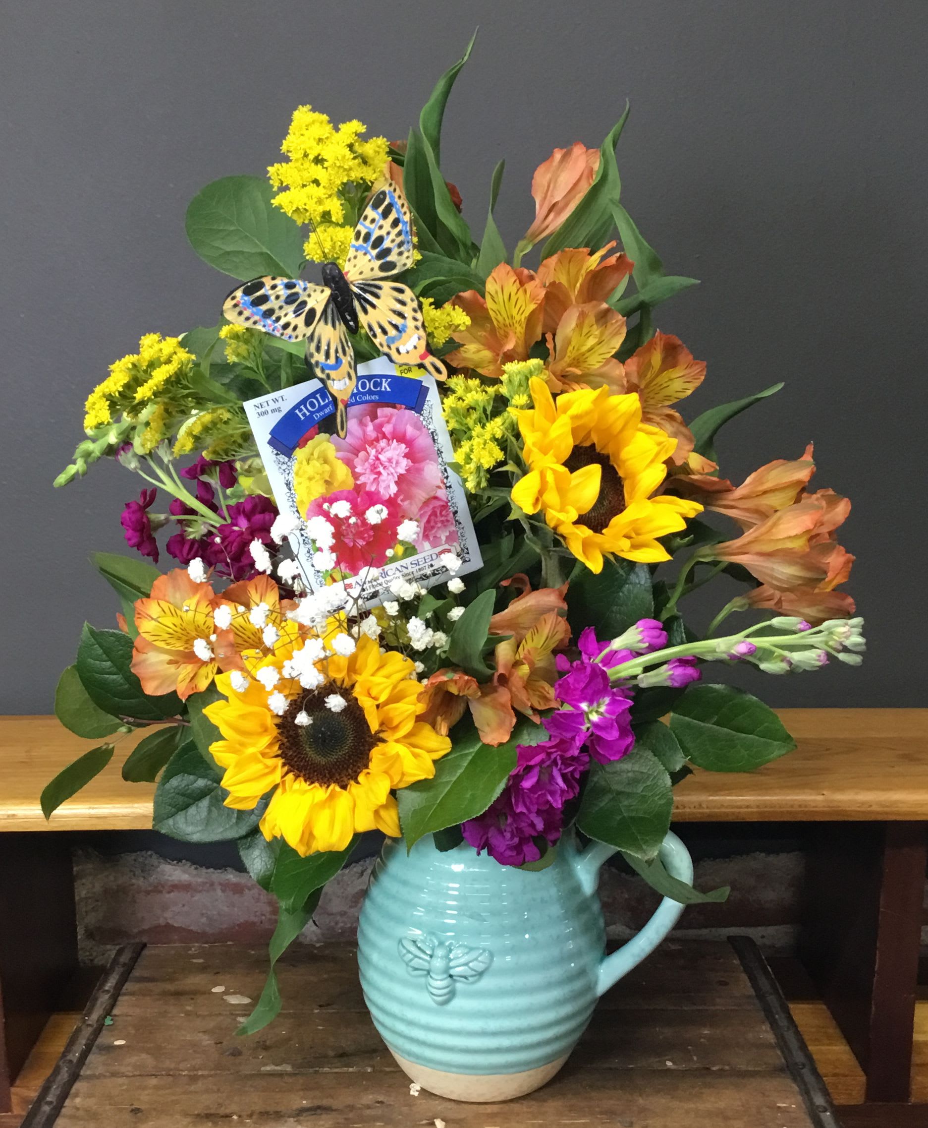 Farmer's Garden - Golden sunflowers and bold orange alstroemeria burst from a light blue ceramic pitcher, making this flower arrangement a joyful choice for a birthday, Mother's Day, or "just because" surprise. The mix of purple snapdragons and delicate white filler blooms creates a lively, garden-fresh palette that reads as both springtime sunshine and warm summer cheer-perfect for saying "thinking of you" or brightening a get-well day.  Presented in a raised-butterfly ceramic pitcher with a decorative butterfly accent and an included seed packet, the Farmer's Garden arrangement feels homemade and hopeful-ideal for grandparents, mothers, or the special woman you're celebrating on Women's Day. The casual, rustic styling and sturdy pitcher make it display-ready for tabletops and kitchen counters; keep blooms in fresh water and trim stems every few days to extend their cheerful life.
