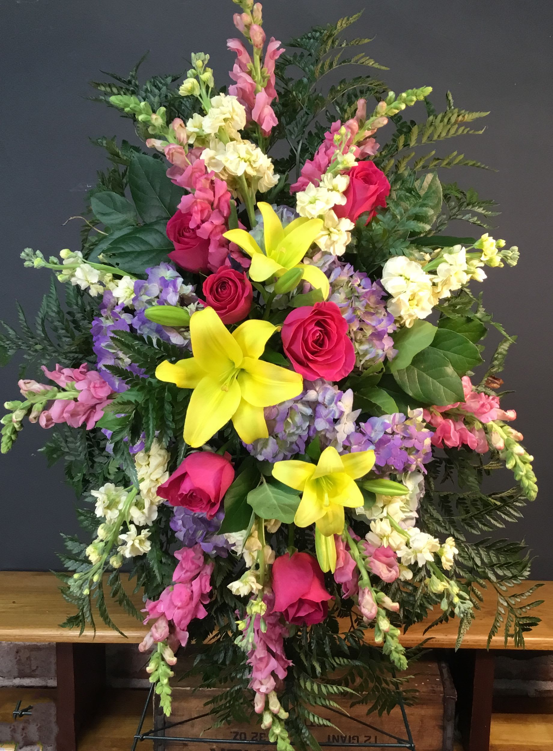Bright and Shining Light Spray - A bold upright spray that greets the room with bright pink roses and sunny yellow lilies, this flower arrangement is a standout choice for celebration or a dignified sympathy tribute. The composition's vibrant color mix-pink, yellow, purple, and soft cream-creates an uplifting yet respectful presence, making it suitable as a birthday or anniversary statement piece or as a thoughtful memorial display to honor a loved one.