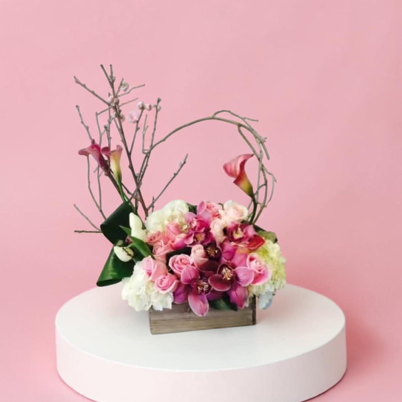 Harmony Bouquet - A sculptural cluster of pink, white, and purple blooms creates a striking centerpiece for weddings, anniversaries, or a special birthday - a romantic flower arrangement that feels both soft and architectural. The Harmony Bouquet pairs elegant calla lilies, classic roses, and graceful orchid-like blooms with looping, curved branches to lift the design and give the person you're celebrating a sense of occasion.  Dense white hydrangea-like clusters form a plush base that contrasts with the richer pinks and magenta tones, while touches of green foliage soften the palette. Arranged in a low wooden box with decorative branches woven through the composition, this piece reads as rustic-modern yet formal enough for a reception table or mantel display.  Perfect for someone who appreciates bold, artistic florals, the Harmony Bouquet makes a memorable statement - she'll feel seen and celebrated. Please allow 2 days lead time on this piece.