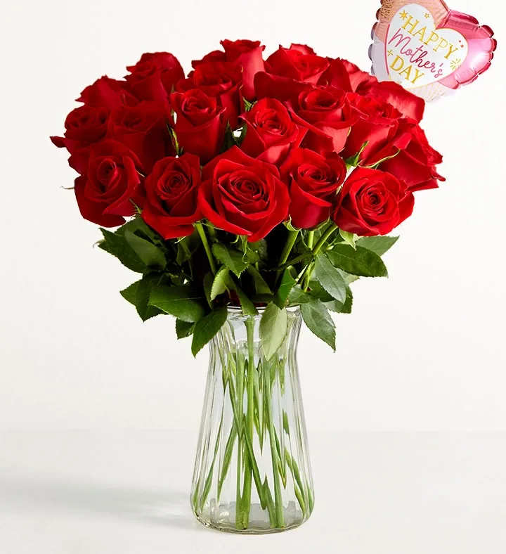 Classic Love Roses  - A full, rounded bouquet of deep red roses set in a clear glass vase-this Classic Love Roses arrangement reads as timeless romance with a Mother's Day twist, perfect as a Mother's Day bouquet, anniversary roses, or a heartfelt "I love you" gift. The dense, tightly packed crimson blooms create an immediate visual impact that will make the person you're thinking of feel cherished and deeply seen.  The arrangement features rich red roses surrounded by fresh green stems and leaves visible through the polished clear glass vase. A pink, heart-shaped foil balloon printed with "Happy Mother's Day" floats above the bouquet, adding a celebratory, family-focused accent that shifts the sentiment toward maternal appreciation without losing its romantic elegance. Colors: red roses, green foliage, and the pink-and-white balloon text combine for a classic, affectionate palette.  This is an ideal choice for honoring a mother, celebrating an anniversary, or sending romantic wishes that also feel warm and familial. The compact, rounded silhouette fits well on a dining table or bedside dresser; vase and balloon arrive ready to display.