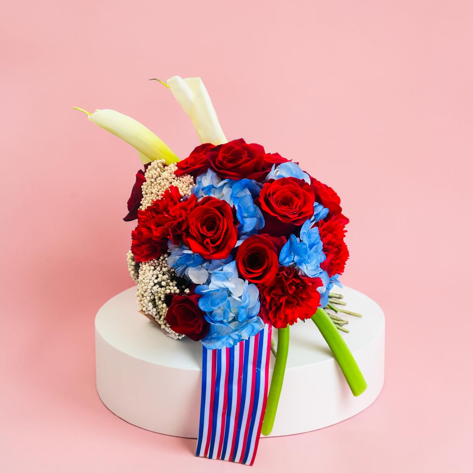 Remembered Always Casket Insert - A dignified casket insert sculpted in a classic cross shape, designed to convey sympathy and honor at a funeral or memorial. The cross's backbone is made from cascading white chrysanthemums that give a soft, reverent texture, while a central sweep of white calla lilies and deep red roses reads as a solemn, devotional focal point-perfect for someone you want remembered always.  Layered green orchid blooms and bright green accents lift the composition and add subtle contrast against the red-and-white palette; touches of yellow and blue appear in the form of hydrangea and small filler clusters for visual depth. The piece is finished with a striped ribbon draping from the base, giving a composed, finished look that photographs well from the casket. Sized and structured as a formal casket spray, it presents as a lasting tribute that will help the person you're honoring feel truly remembered.