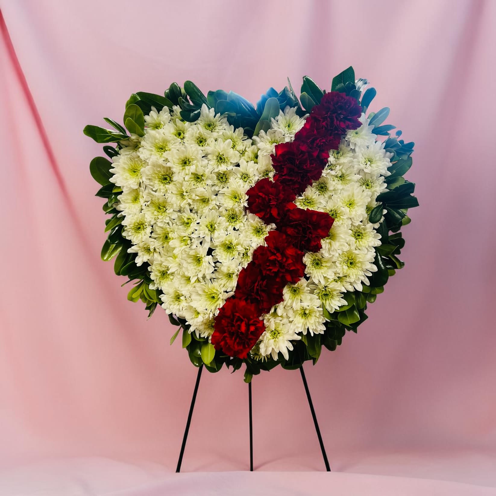Broken Heart Spray - A heart-shaped floral tribute that reads as a quiet, heartfelt message-perfect for sympathy and funeral services or for sending a sincere "I'm thinking of you." This Broken Heart spray pairs delicate white daisy-like blooms with a bold diagonal band of deep red carnations, framed by rich green foliage and touched with subtle blue accents. For birthdays, Mother's Day, or a "just because" moment, it also serves as a respectful way to mark a meaningful milestone or offer comfort.  The piece is shown on a sturdy metal easel stand for dignified display and is crafted with dense, evenly spaced flowers to maintain its defined heart shape. The white daisies provide a soft, comforting texture while the red carnations create a focused, emotional stripe; dark greenery around the edges gives the spray visual weight and contrast. Shown both as a rounded white-rose vase arrangement and as the heart-shaped easel spray, the design emphasizes clean lines and classic sympathy palette.  Choose this arrangement when you want the recipient to feel truly seen-whether you're offering condolences, saying thank you, congratulating someone in a formal way, or celebrating a birthday with quiet elegance. The easel display makes it simple for funeral placement; if you need delivery details or size options, we'll help confirm availability and arrange timing to suit the service or event.