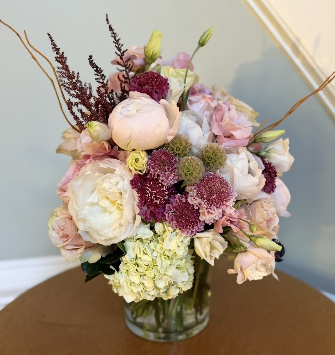 Pastel bouquet of roses, peonies, hydrangea, and purple accent flowers in a glass vase