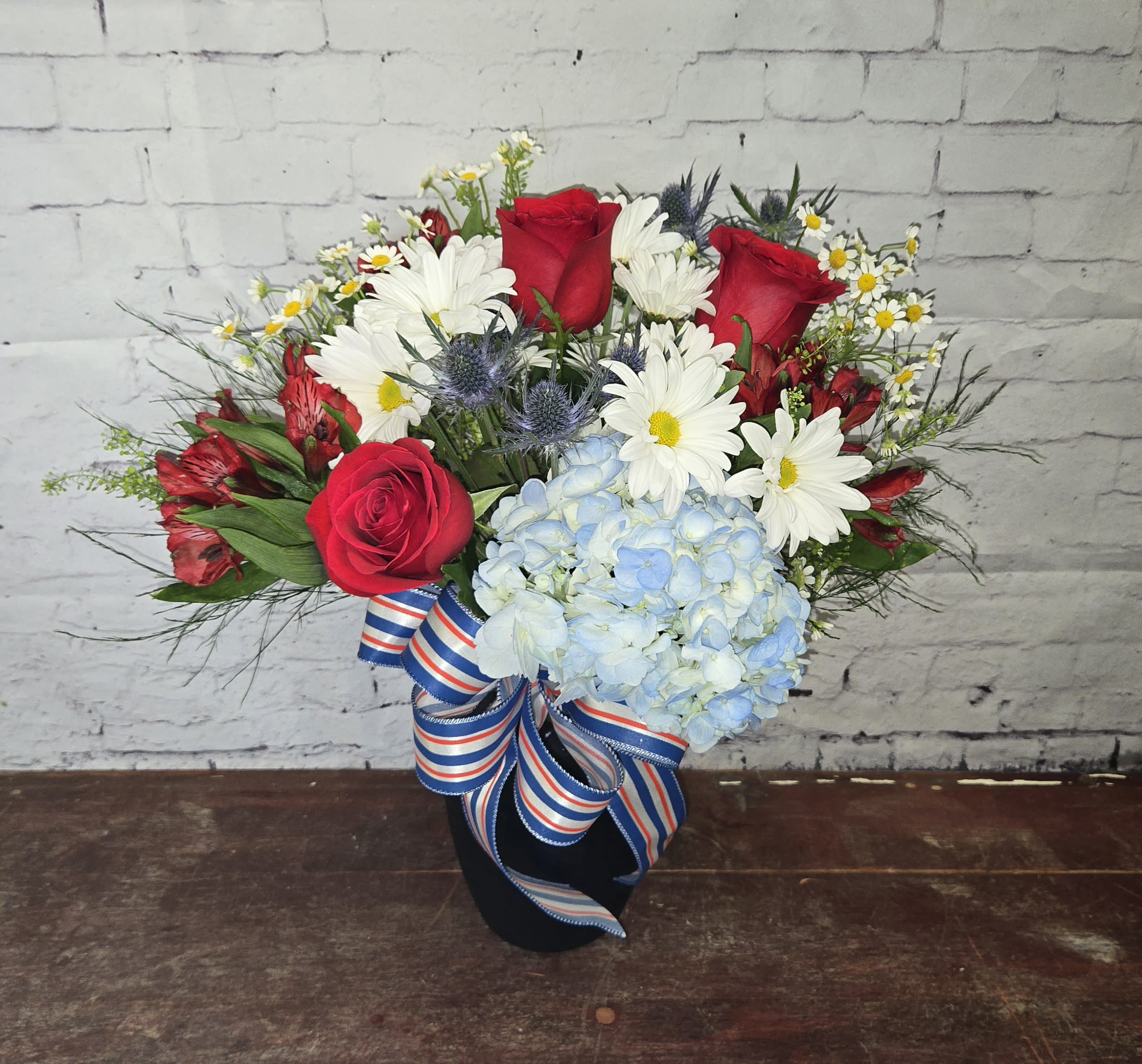 Bouquet of red roses, white daisies, and blue hydrangea in a black vase