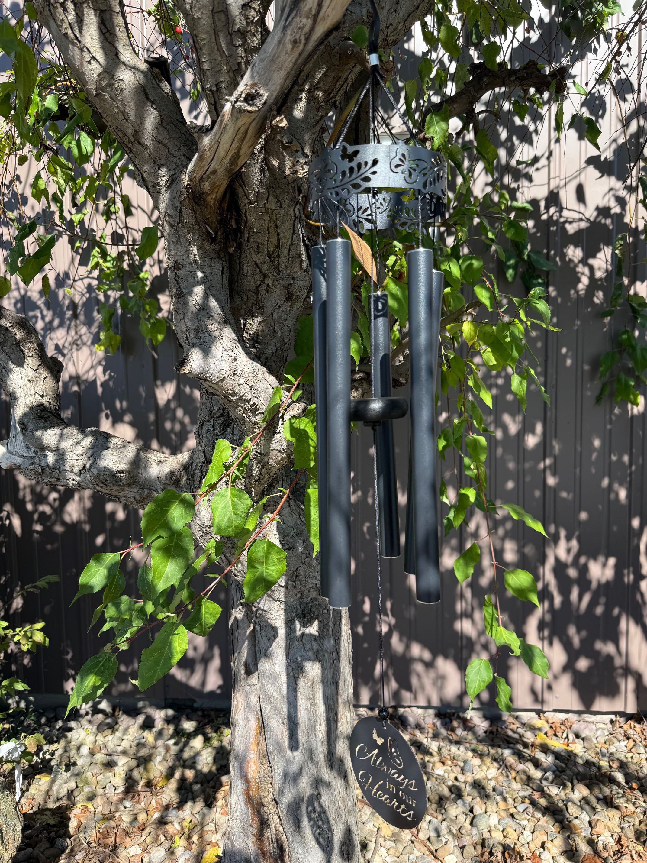 Black metal wind chime with memorial message hanging from a tree outdoors.