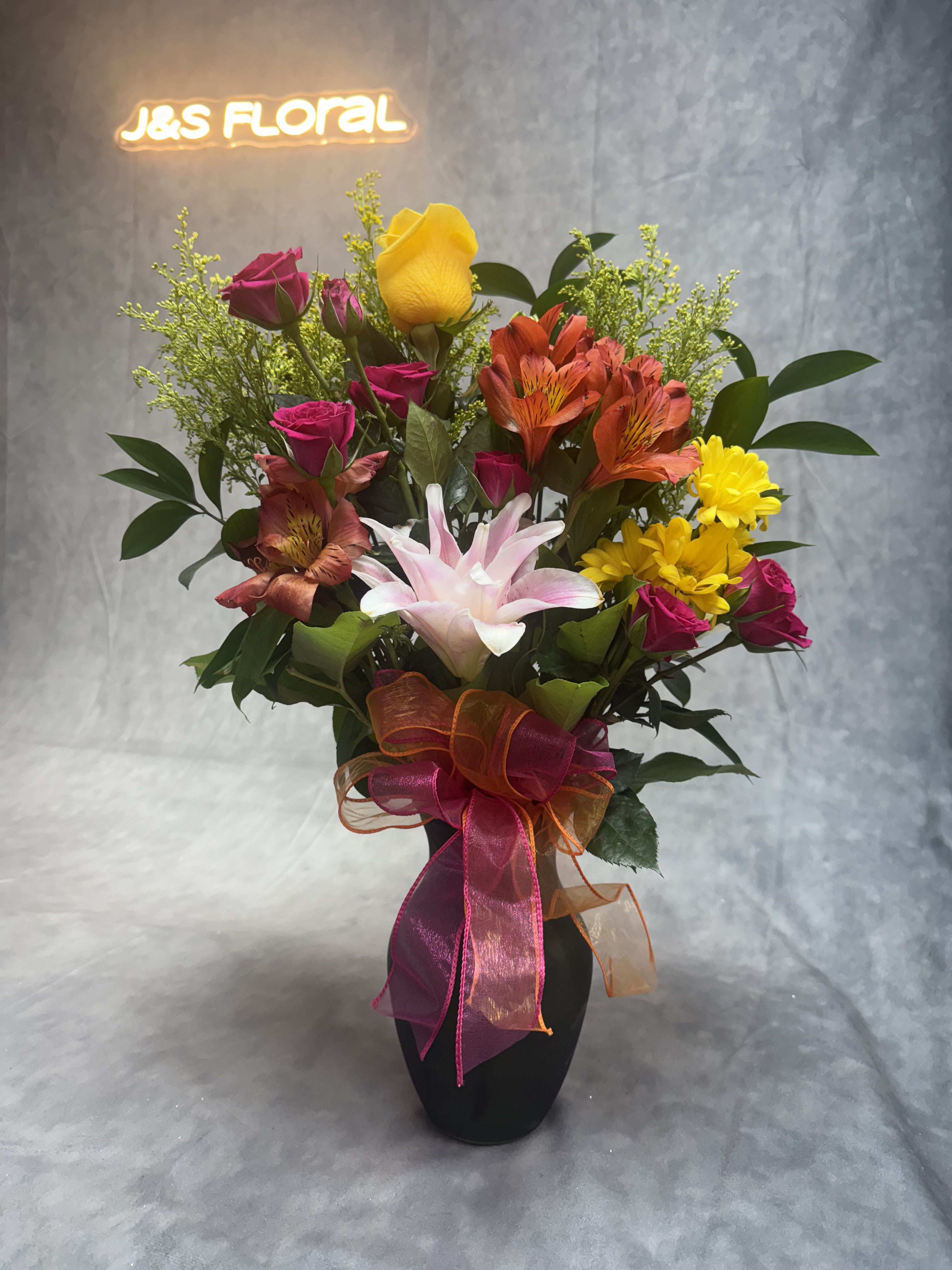 Mixed bouquet of roses, lilies, and daisies in a black vase with ribbon