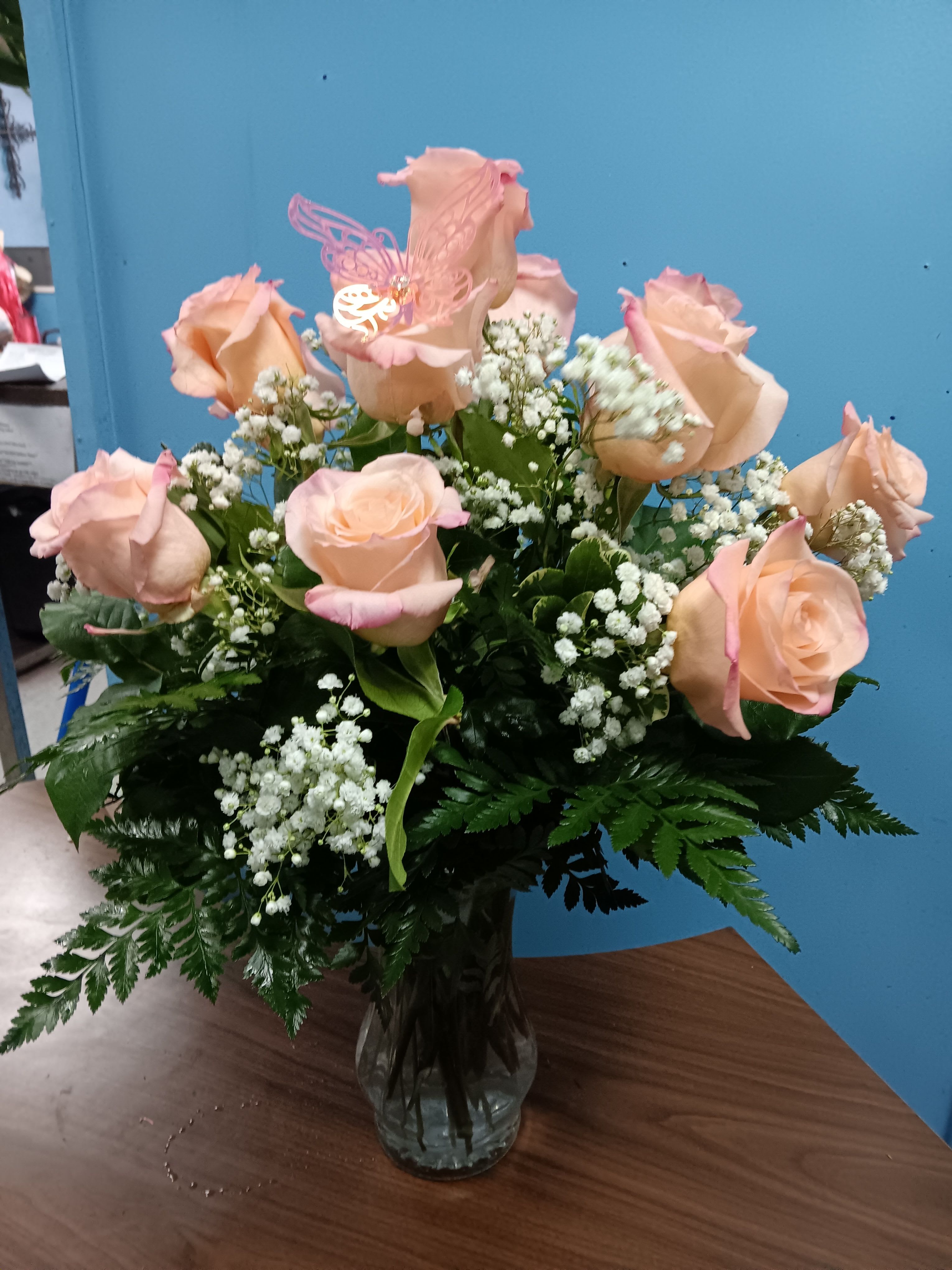 1Dz Pink Roses - Soft peach-pink roses gathered in a rounded, upright bouquet create a tender, romantic gift perfect for Mother's Day, anniversaries, birthdays, or simply to say "I'm thinking of you." This dozen-rose arrangement brings a gentle, uplifting mood with closely grouped blooms and a playful pink butterfly pick tucked into the center for a whimsical touch that catches the eye.  Each arrangement pairs the peach roses with delicate white filler flowers and a dense base of greenery, arranged in a clear glass vase for a classic presentation that fits any room. The layered petals and soft pink-tinged edges add texture and depth, while the butterfly accent adds personality. Available in standard dozen sizing (with larger deluxe and premium options noted), it's an ideal choice for get well, new baby, thank you, sympathy, graduation, housewarming, or just-because moments-seasonally sweet for Easter yet timeless enough for spring, summer, autumn, or winter gifting.