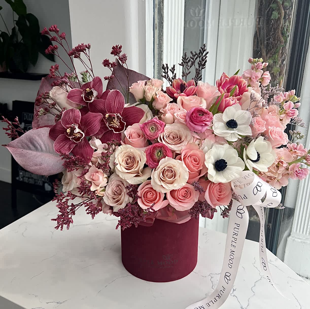 Pink and burgundy floral arrangement with roses, orchids, and white anemones in a round velvet hatbox.
