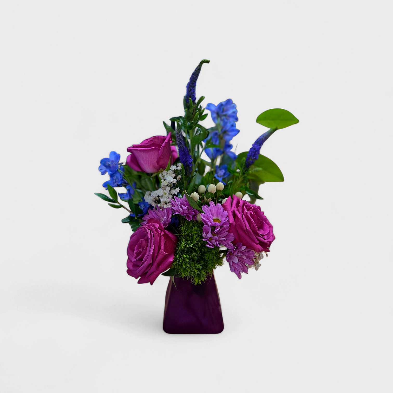 Whole Heartedly - Vivid magenta roses take center stage in this Mom purple flower arrangement, a bright and cheerful bouquet perfect for birthdays, Mother's Day, or a "thinking of you" surprise. The mix reads jewel-toned - purple, blue, pink, and white - so the person you're sending it to will feel celebrated by a bold, happy splash of color rather than a timid nod.  A compact base of small purple daisy-like blooms and white accents gives texture while taller blue spike flowers lift the design into a lively, slightly asymmetrical silhouette. Arranged in a purple glass vase, the saturated palette feels cohesive and decorative; the vase both anchors the composition and makes the gift ready to display.  This arrangement suits everyday gifting or special moments when you want her to feel genuinely seen and uplifted. Care is simple: keep fresh water in the vase, trim stems occasionally, and place out of direct heat to extend bloom life.