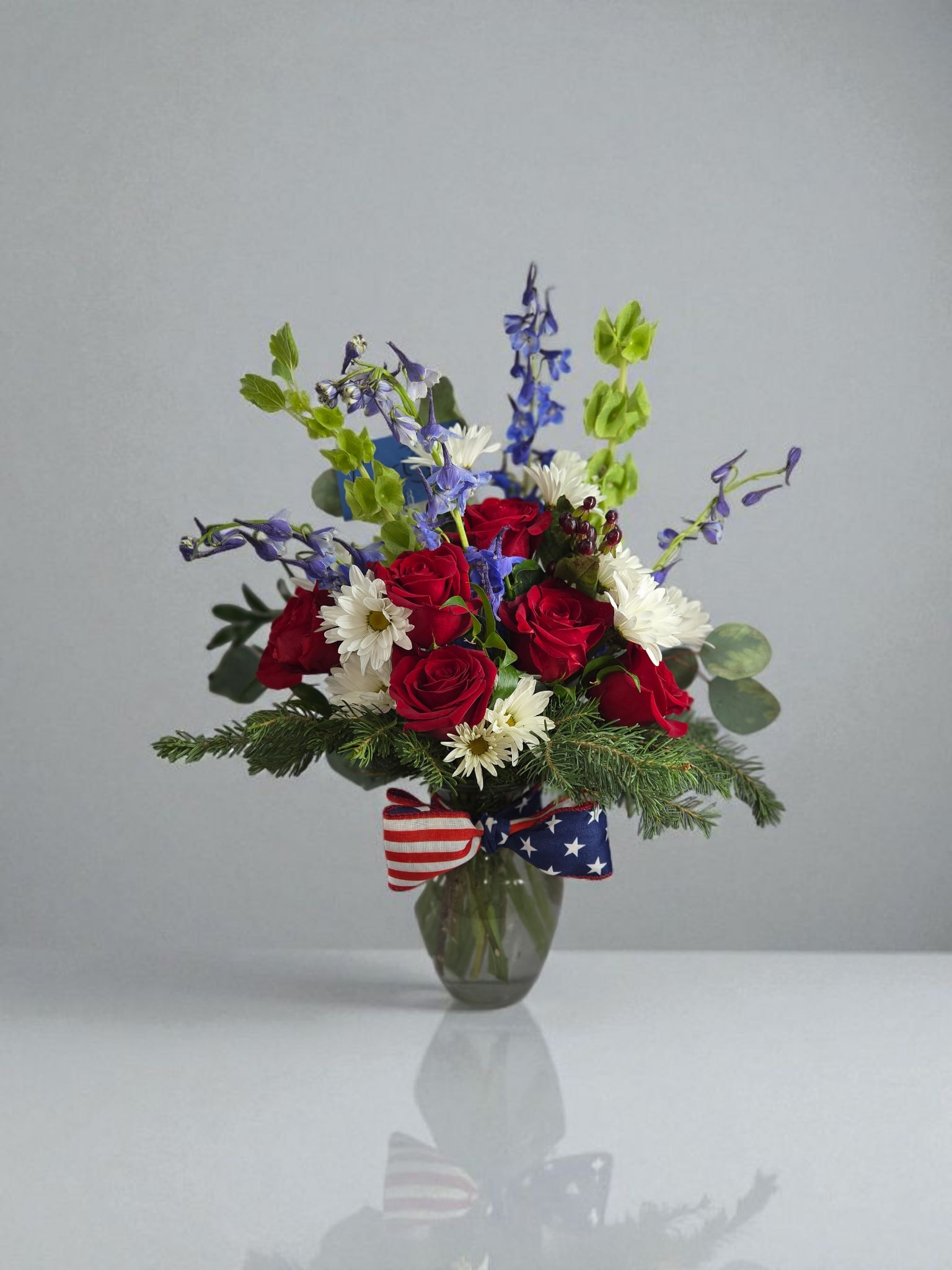 Red roses and white daisies in a glass vase with a patriotic ribbon