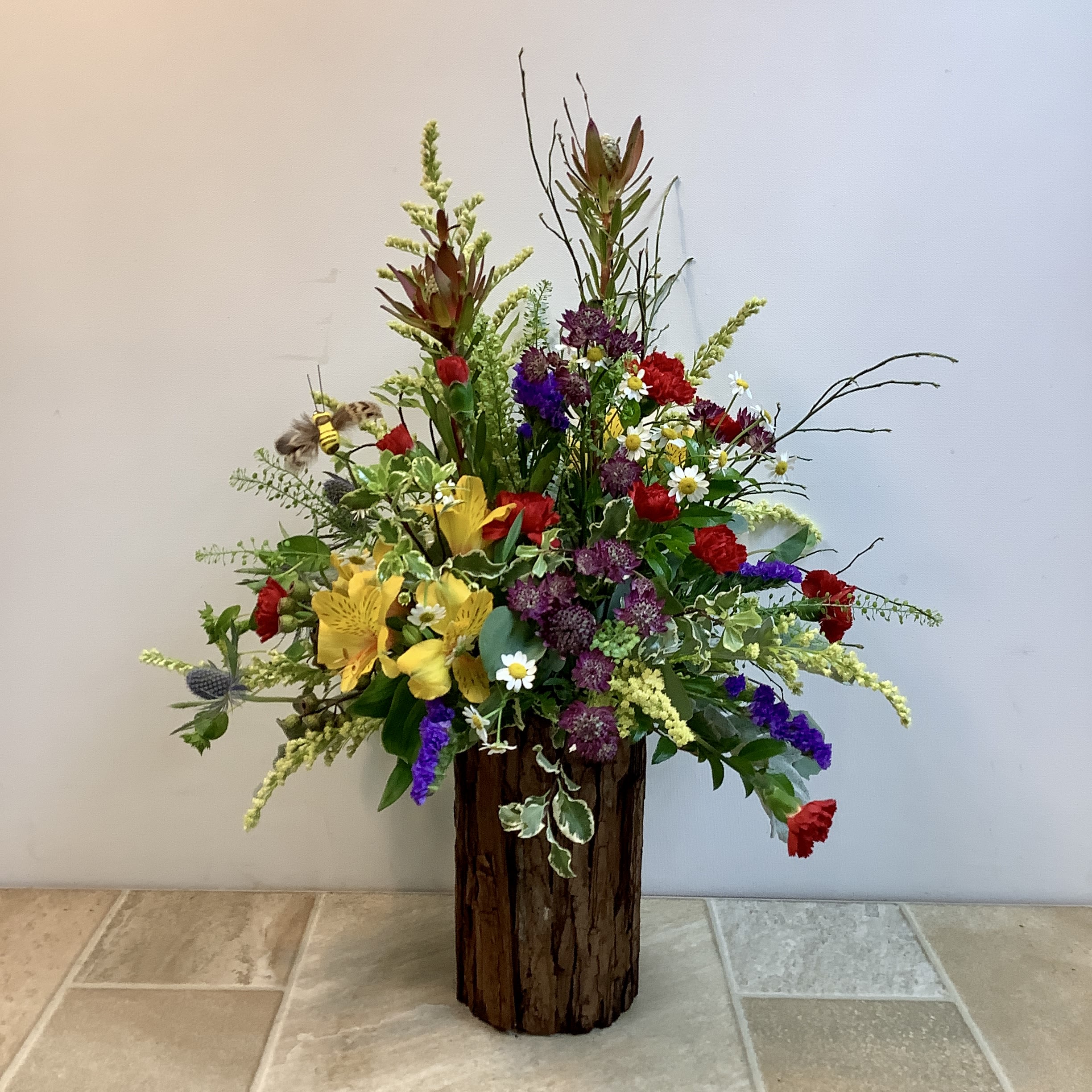 Woodland Wilds - This large arrangement has a lively, meadow-inspired feel with a slightly rustic, woodland twist. It’s designed in a natural wood cylinder vase, which grounds the piece with earthy texture while letting the flowers feel fresh and untamed. Bright yellow alstroemeria create a sunny focal point, surrounded by pops of red, purple blooms. Airy sprigs of greenery and delicate filler flowers weave throughout, giving it that “just gathered from the field” look. A small decorative bee accent enhances the cheerful, pollinator-friendly vibe.  Approximate size: 16" w x 23" h  Primary flowers: lily, carnation