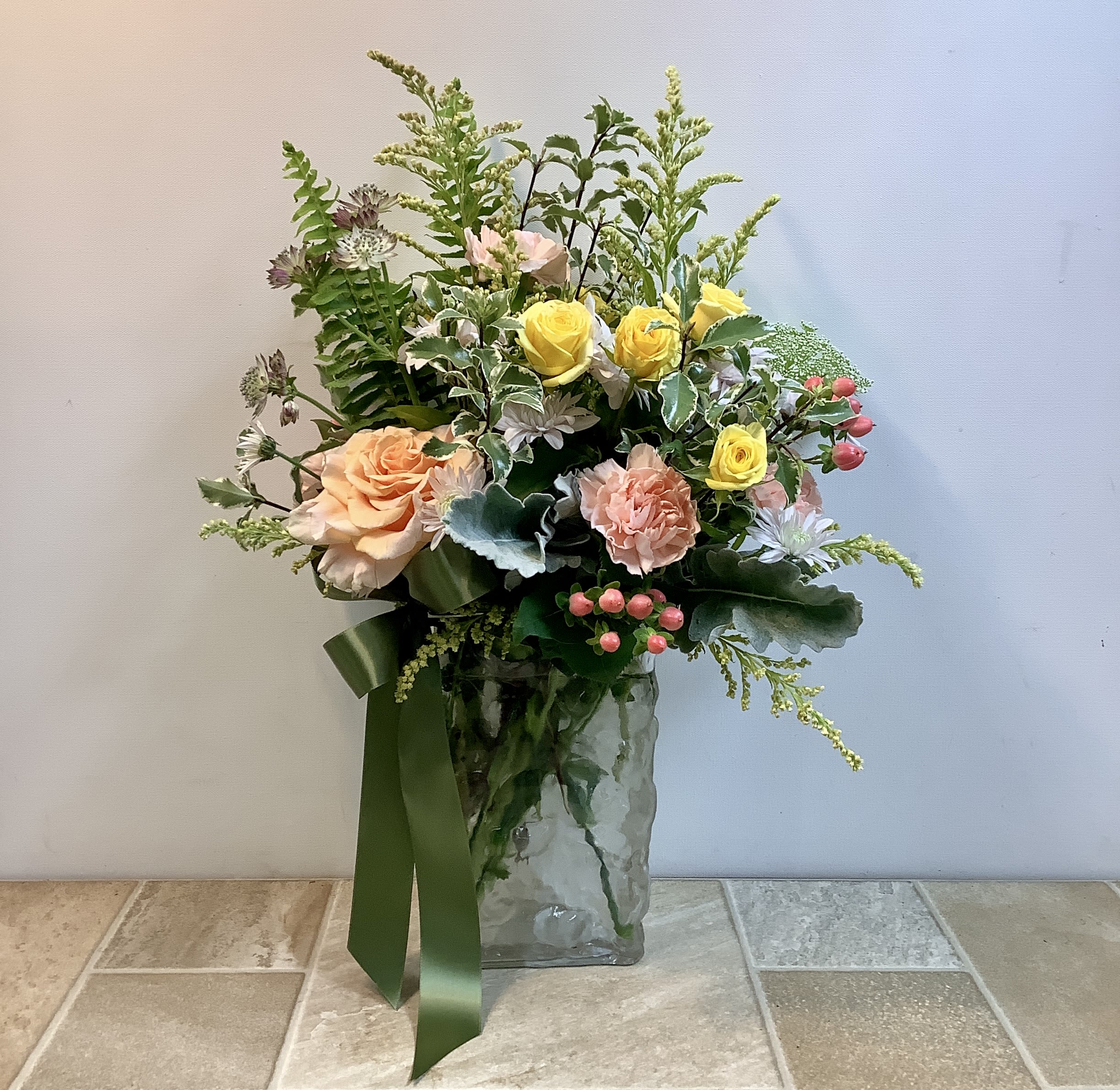 Peachy Keen - This arrangement has a soft, garden-inspired feel with a natural, airy structure. It features a cheerful mix of warm peach and sunny yellow roses, paired with peachy-pink carnations, delicate accent blooms, and pops of coral-pink berries. The flowers are arranged in a clear, textured glass vase and finished with a wide green ribbon for a polished, gift-ready feel.  Approximate size: 15" w x 23" h  Primary flowers: rose, carnation, mum