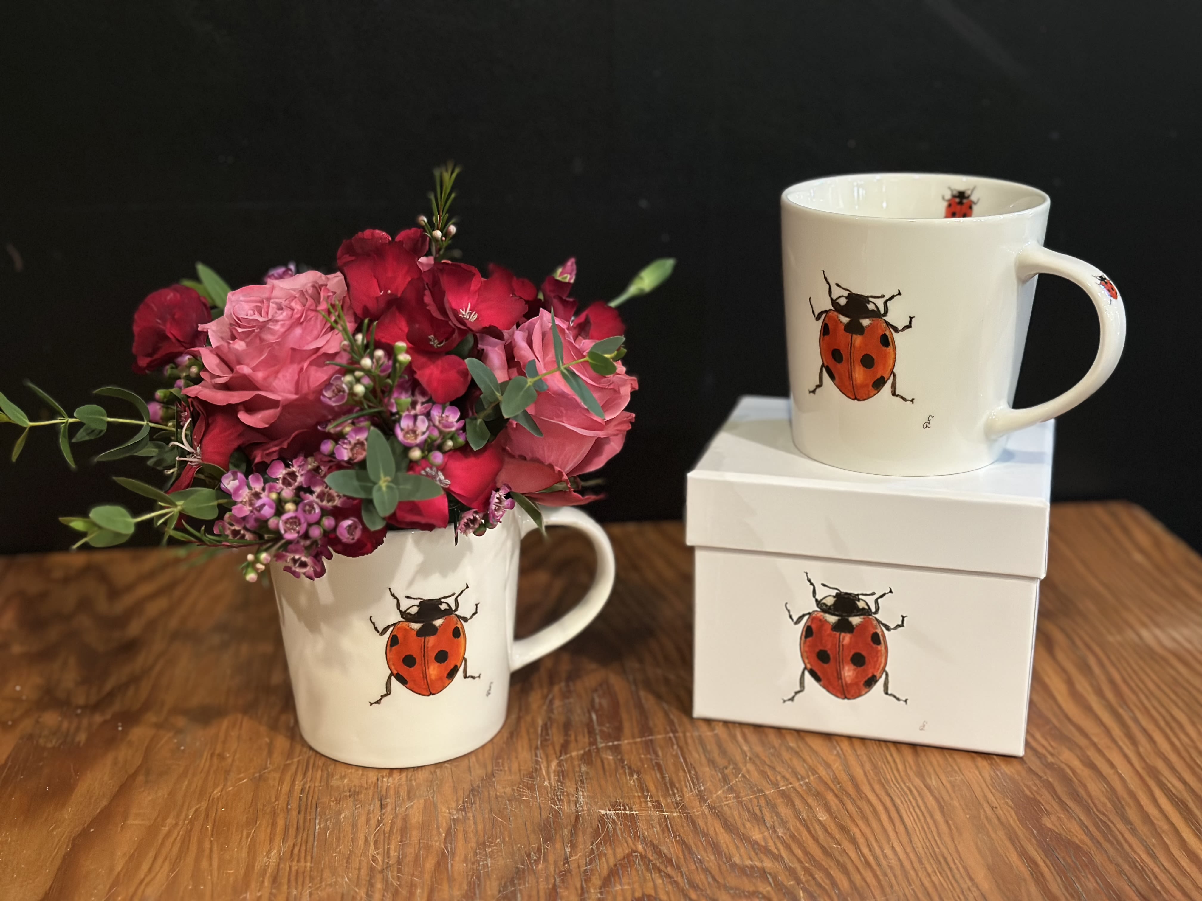 Pink and red flower bouquet in a ladybug mug beside matching gift boxes