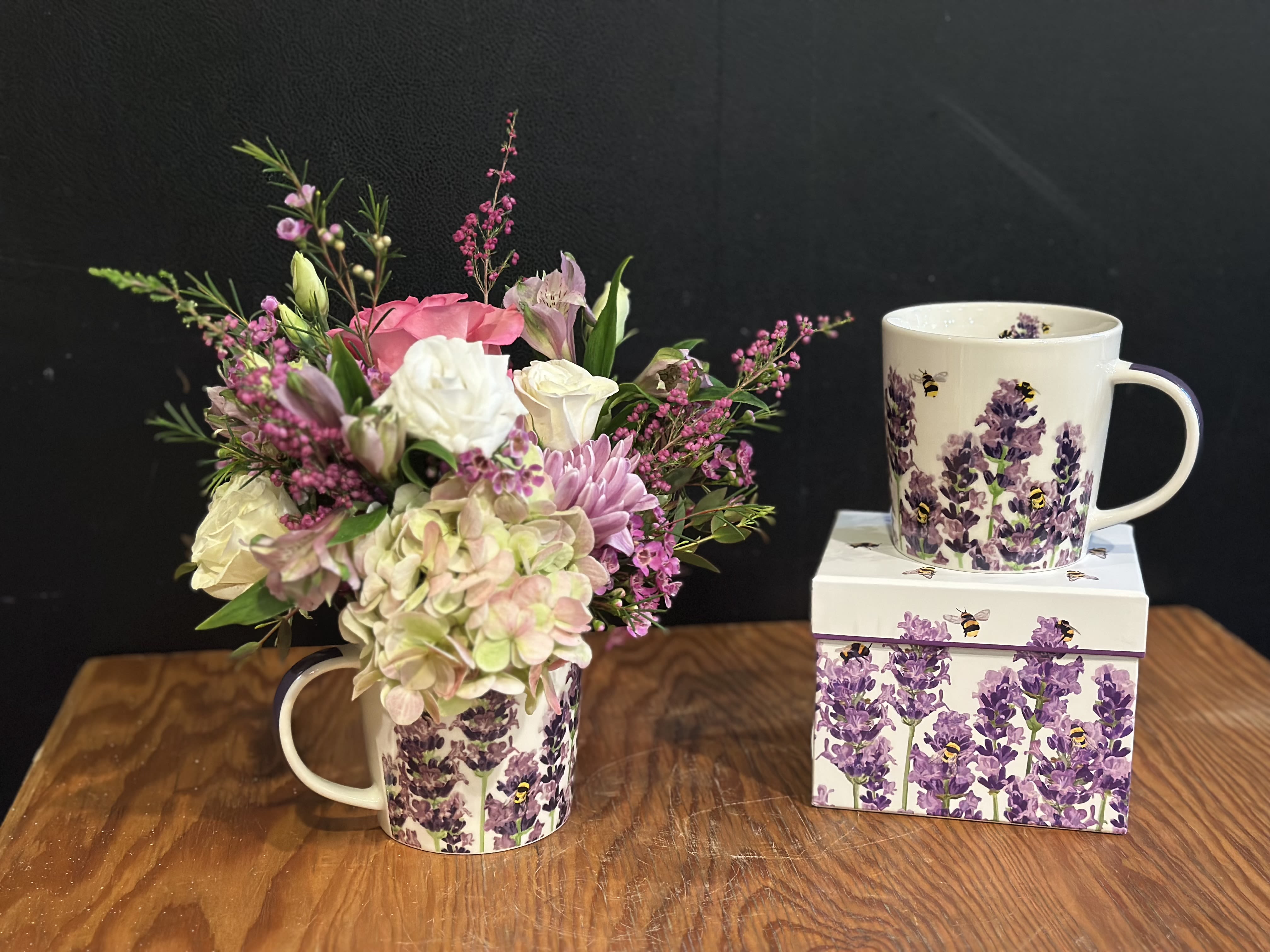 Pink and white flower arrangement in a lavender bee-patterned mug beside a matching mug and box