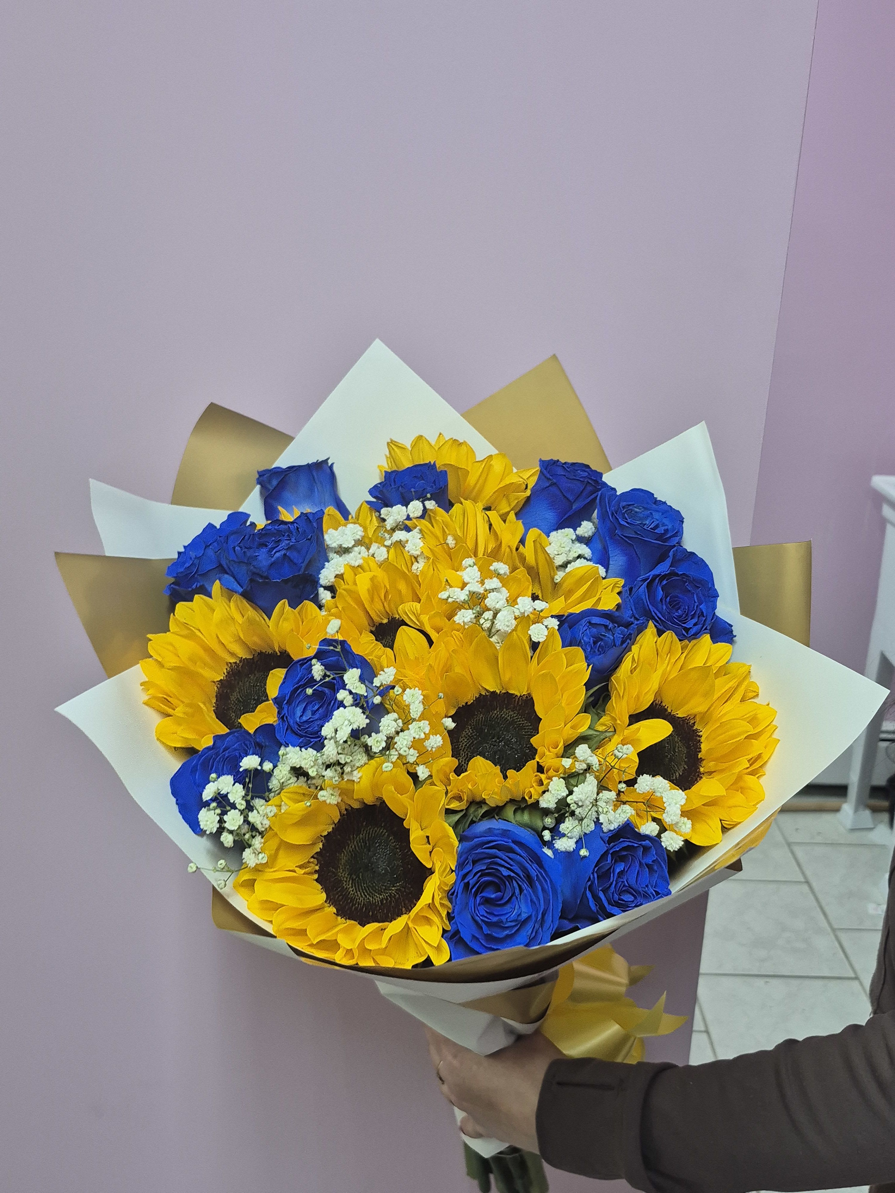 Radiant Blue Medley - The Radiant Blue Medley is a bold and beautiful bouquet that pairs bright yellow sunflowers with eye catching blue roses. Accented with tiny white flowers for a soft touch, this arrangement creates a stunning contrast of warm and cool colors. Wrapped in crisp white and gold paper, it is a simple yet striking gift that brings a modern, cheerful energy to any room.