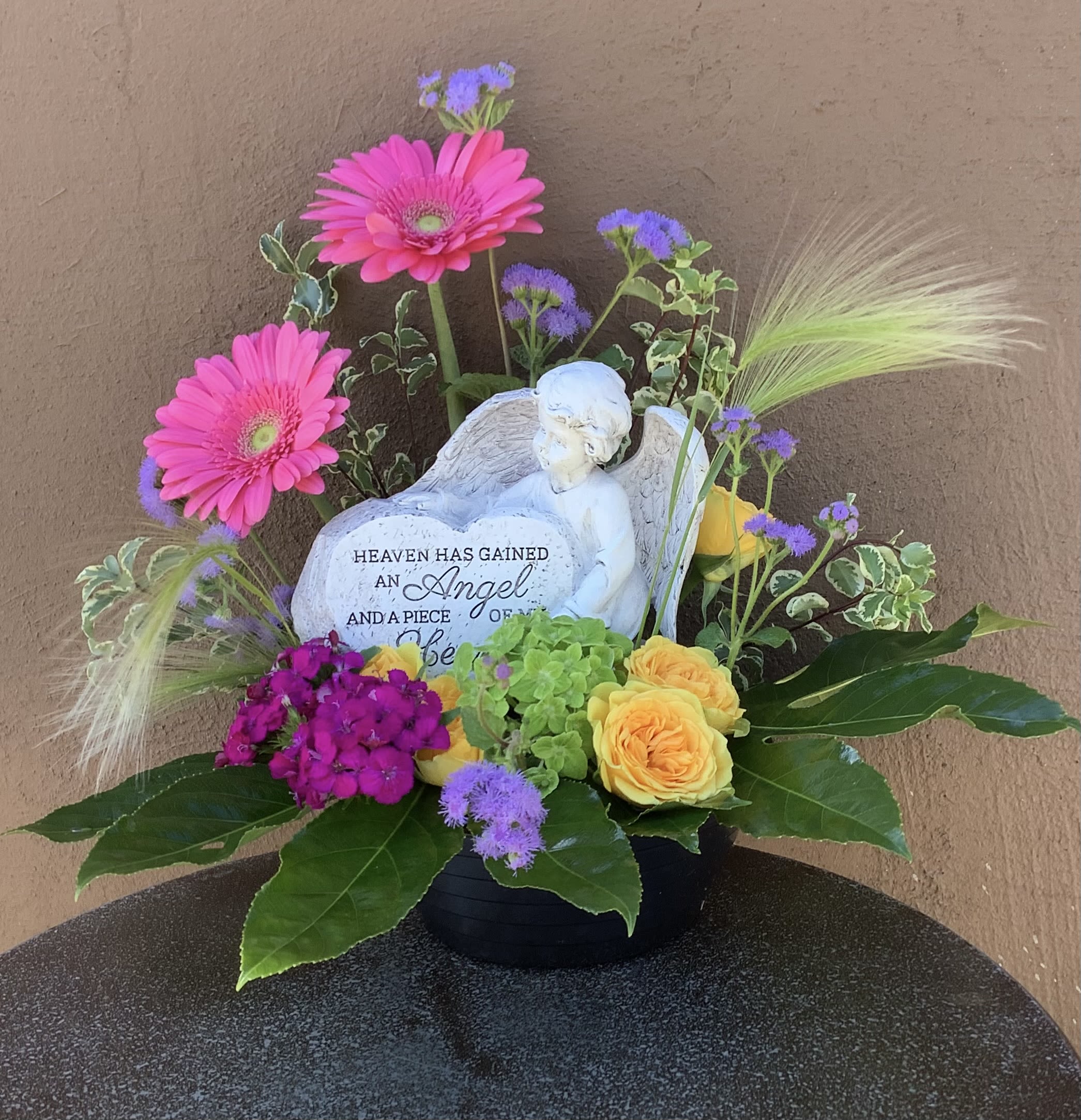 Floral sympathy arrangement with pink gerbera daisies and yellow roses around an angel figurine