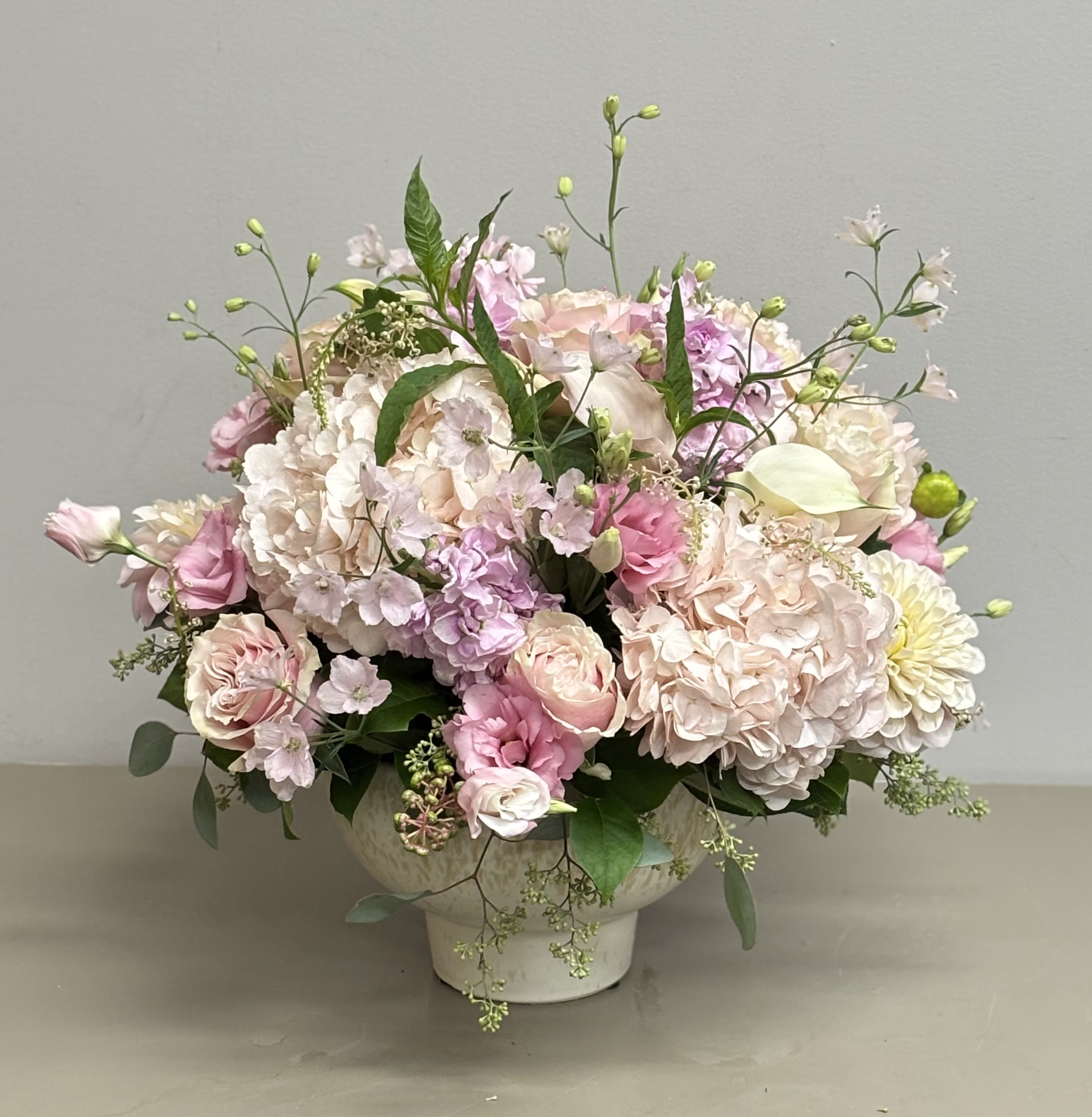 Low arrangement of blush and pink roses, hydrangeas, and mixed blooms in a white pedestal bowl