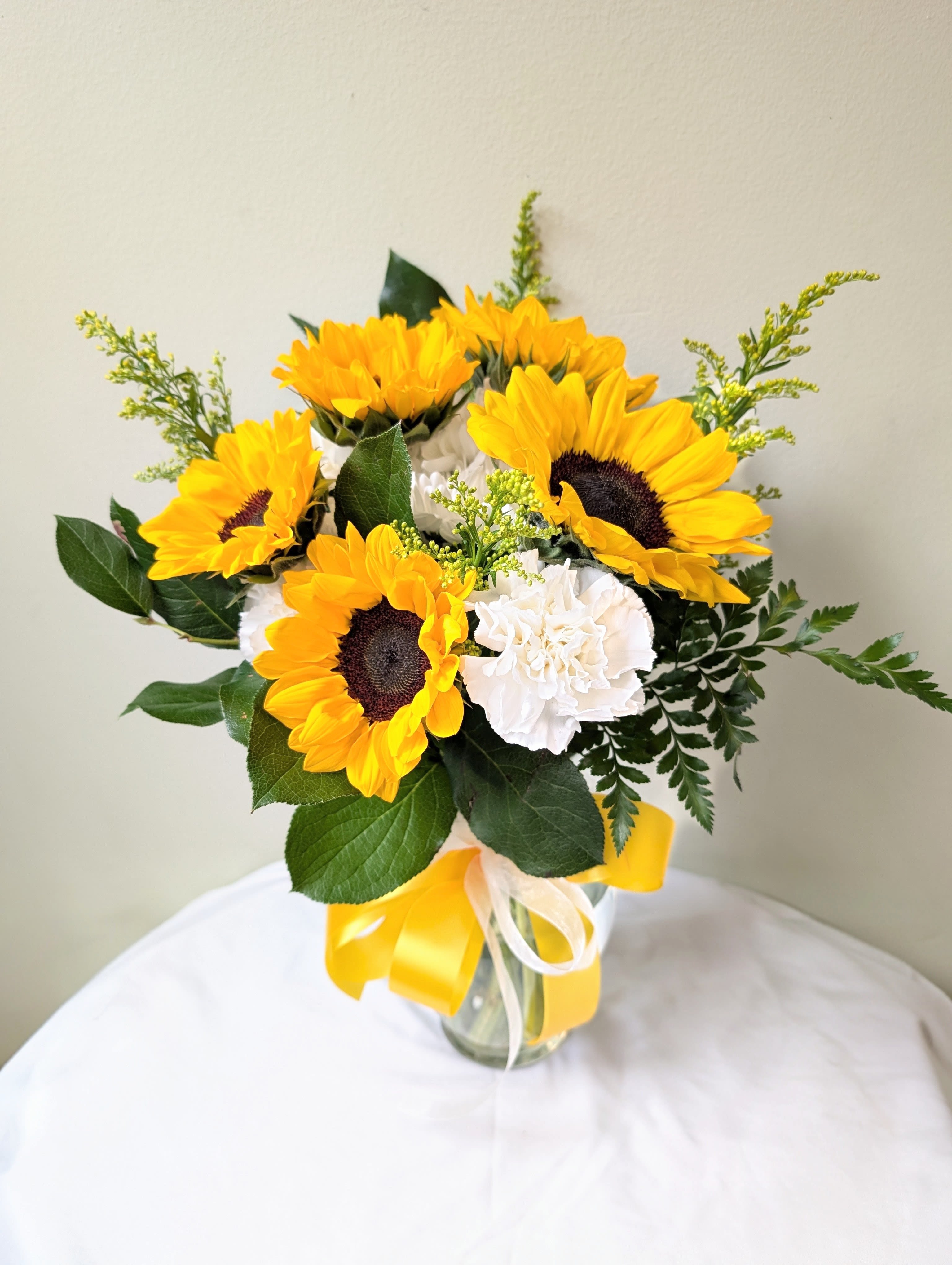 Pure Sunflowers - Bright, cheerful sunflowers take center stage in this Pure Sunflowers arrangement, made for birthdays, thank-you gestures, or as a bright get-well pick-me-up. The bold yellow blooms paired with a single white carnation accent create a fresh, high-contrast look that makes the recipient feel genuinely seen and uplifted-perfect for the person who needs a little sunshine in their day.  Arranged in a clear glass vase and finished with a wide yellow ribbon plus a delicate white tie, this compact bouquet includes dark green foliage and airy filler stems for a slightly rustic, full appearance. The glass vase lets the stems show through for a clean, modern touch; no special care beyond fresh water and a cool spot is needed to keep the flowers looking their best for several days. Order this as an everyday gift that sends a warm, optimistic message.