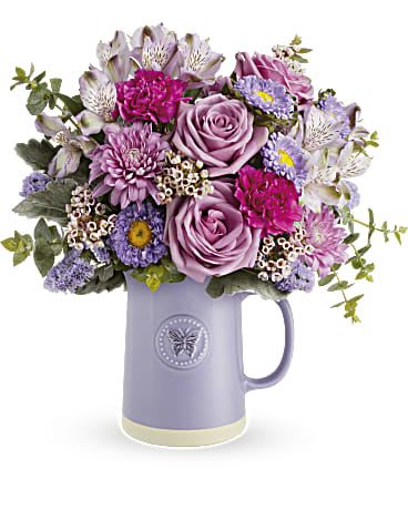Sweetest Flutter - Lavender and soft pink roses take center stage in this garden-fresh flower arrangement, arranged in a feminine lavender ceramic pitcher vase that's perfect for a birthday, Mother's Day, anniversary, or a thoughtful "just because" surprise. The Sweetest Flutter bouquet reads as romantic and airy-she'll immediately notice the soft color wash and the relaxed, rounded silhouette that feels like a handpicked posy from a spring garden.  Bright magenta carnations and pale alstroemeria add lively contrast while small purple aster blooms and lavender chrysanthemums bring delicate texture and movement among green foliage. The ceramic pitcher makes a charming keepsake vase that's ready to display on a kitchen table or bedside, approximately medium-sized for everyday gifting. Designed to make the recipient feel genuinely seen-whether you're saying thank you, celebrating a birthday, or simply thinking of them-this arrangement arrives ready to enjoy with simple care: fresh water and a cool spot away from direct sun will help blooms last.