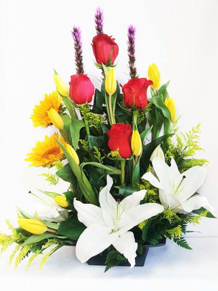 Tall mixed arrangement of white lilies, red roses, yellow tulips and sunflowers in a low black container