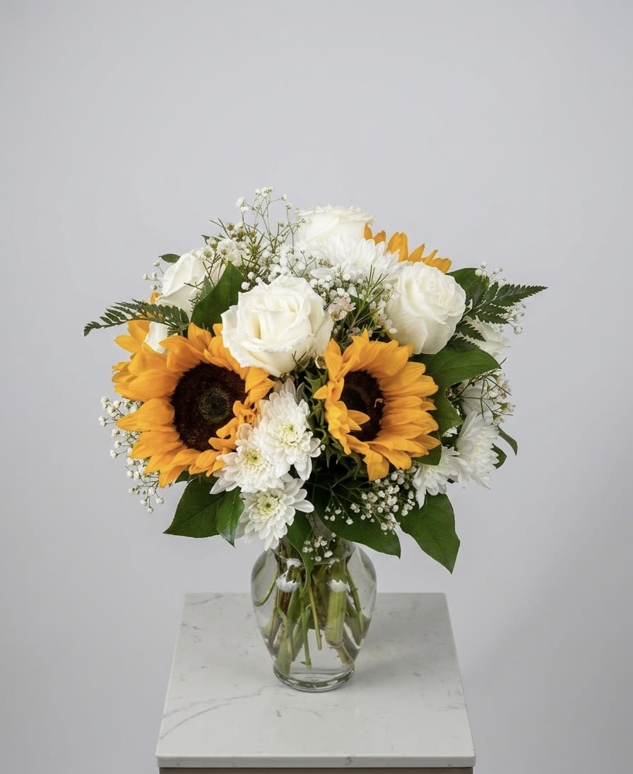 The  Sunburst Classic - Elevate your space with The Classic Dawn, an exclusive artisanal floral design from Lovely Flowers Boutique. This signature vase flower arrangement features the vibrant warmth of sunflowers paired with the timeless elegance of premium white roses. Unlike standard bouquets, our professional florist team hand-styles these blooms in a glass vase for a polished, "studio-ready" look. It’s the perfect classic choice for Mother’s Day, corporate gifts, or sophisticated home decor.