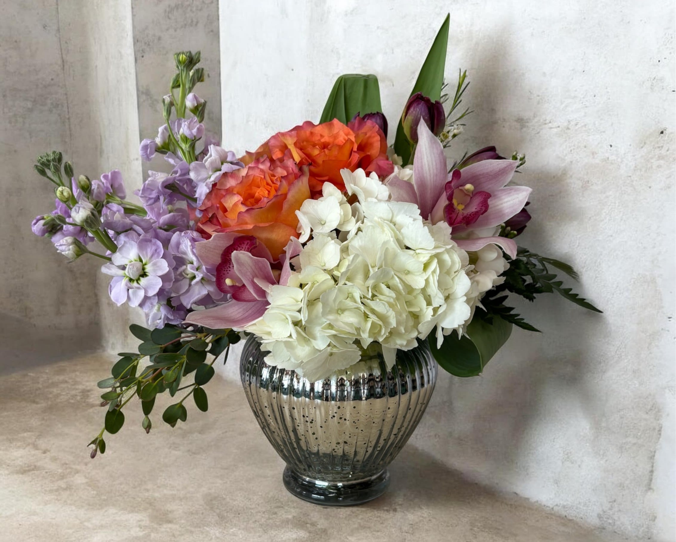Grey House Blooms-Delight - Round, lush mix of white hydrangea, orange roses, pink orchids, tulips and lavender stock in a silver mercury vase.