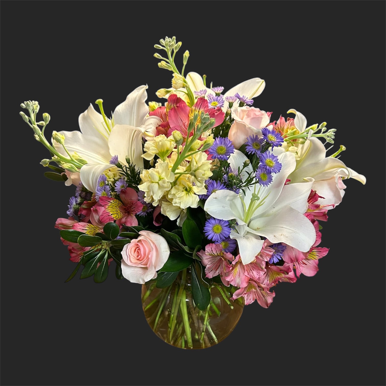 A Sunday Brunch  - Large white lilies open wide among soft pink roses to create a cheerful, slightly romantic flower arrangement perfect for a birthday bouquet, anniversary flowers, or a thoughtful thank-you gift. The mix of pink alstroemeria and small purple aster-like blooms gives a lively, textured contrast while pale yellow accents warm the palette-making the person you're sending this to feel noticed and truly celebrated.  Arranged in a classic rounded glass vase, the composition is full and balanced with blooms fanning outward in an abundant, relaxed cluster. Expect fragrant white lilies, velvety pink roses, delicate pink alstroemeria, and clusters of purple daisy-style asters with pale yellow filler for contrast; the vase keeps it elegant and easy to display. Ideal for a bedside table, dining centerpiece, or office desk-no special care beyond fresh water every couple of days will keep the bouquet looking fresh for as long as possible.