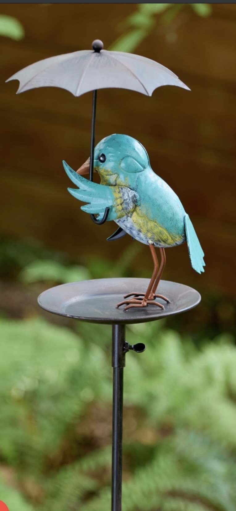 Rainy Day Bird Feeder - A whimsical blue bird clutching a tiny umbrella perches above a round feeder tray-this Rainy Day Bird Feeder is a playful garden stake that brings charm to any outdoor space. As a "just because" gift or thoughtful housewarming or birthday surprise, it creates a small, joyful moment for the person you're celebrating and invites neighborhood birds to investigate.  Crafted with a painted metal bird in turquoise, yellow, white and brown accents, the feeder sits on a sturdy black metal stake and tray that withstands garden weather while keeping a handcrafted, rustic look. The umbrella detail and compact tray make it ideal for seed offerings or decorative display in a flower bed, container garden, or by a patio. Easy to place in soil and low-maintenance, it's the kind of gift that lets someone know you're thinking of them without fuss.