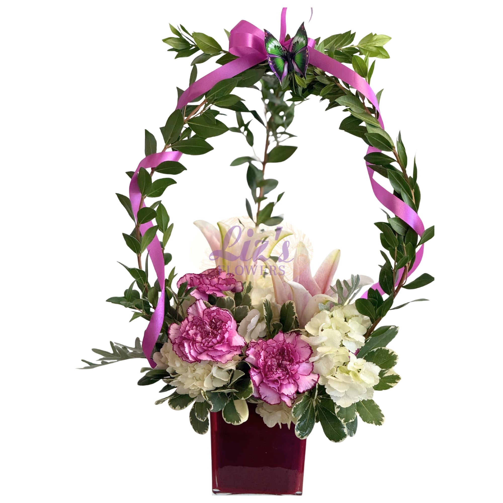 Enchanted Garden - Whimsical and artistic design featuring an elegant arch of lush greenery that frames a delicate cluster of purple Carnations, white Hydrangeas, and soft pink Lilies. Accented with a flowing lavender ribbon and a playful butterfly detail.