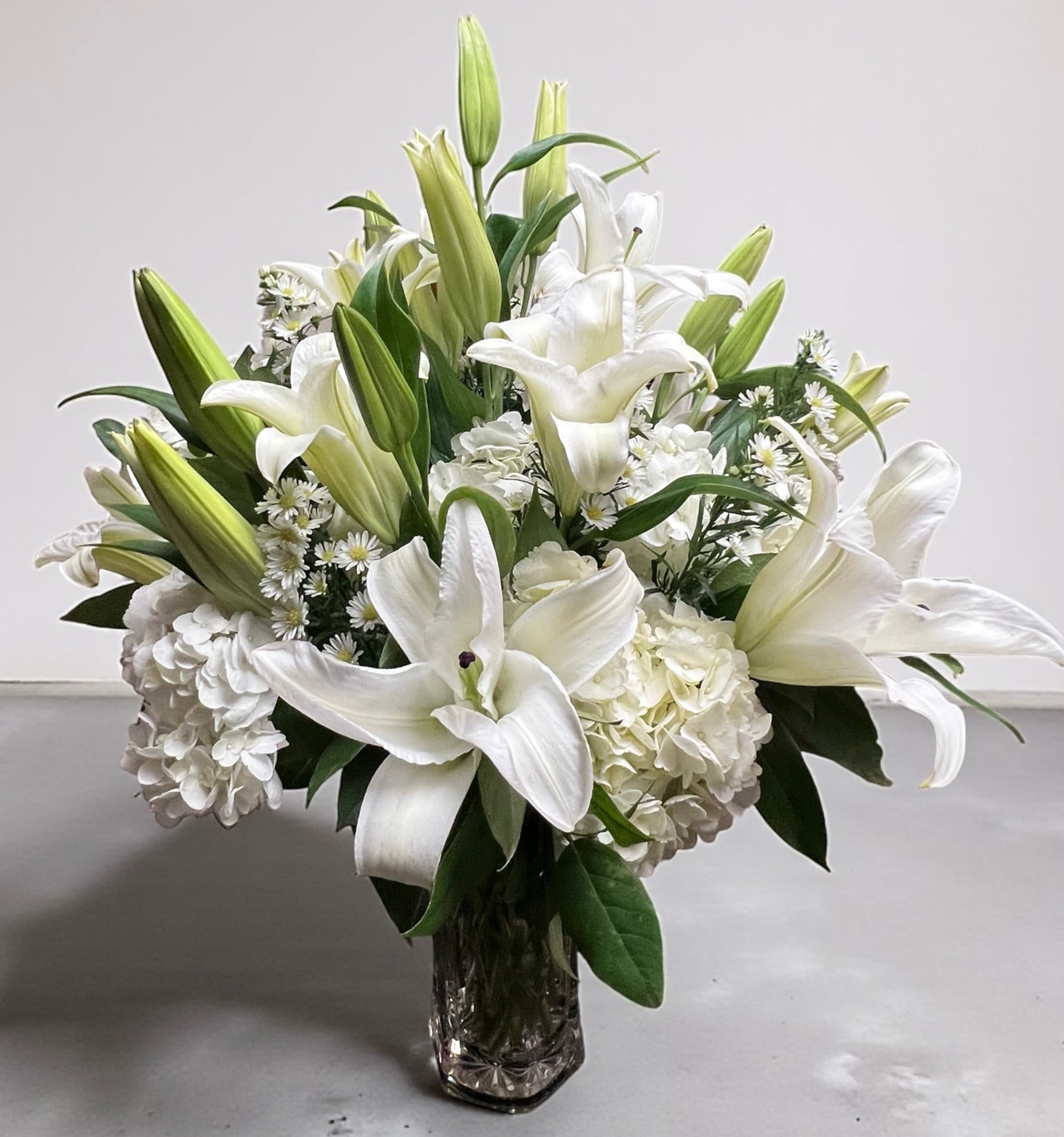 Ivory Embrace - Graceful, serene, and timeless- Ivory Embrace is a breathtaking all white arrangement designed to bring comfort, beauty, and peace to any space. Featuring elegant white lilies, lush hydrangeas delicate accent florals nestled in rich greenery.