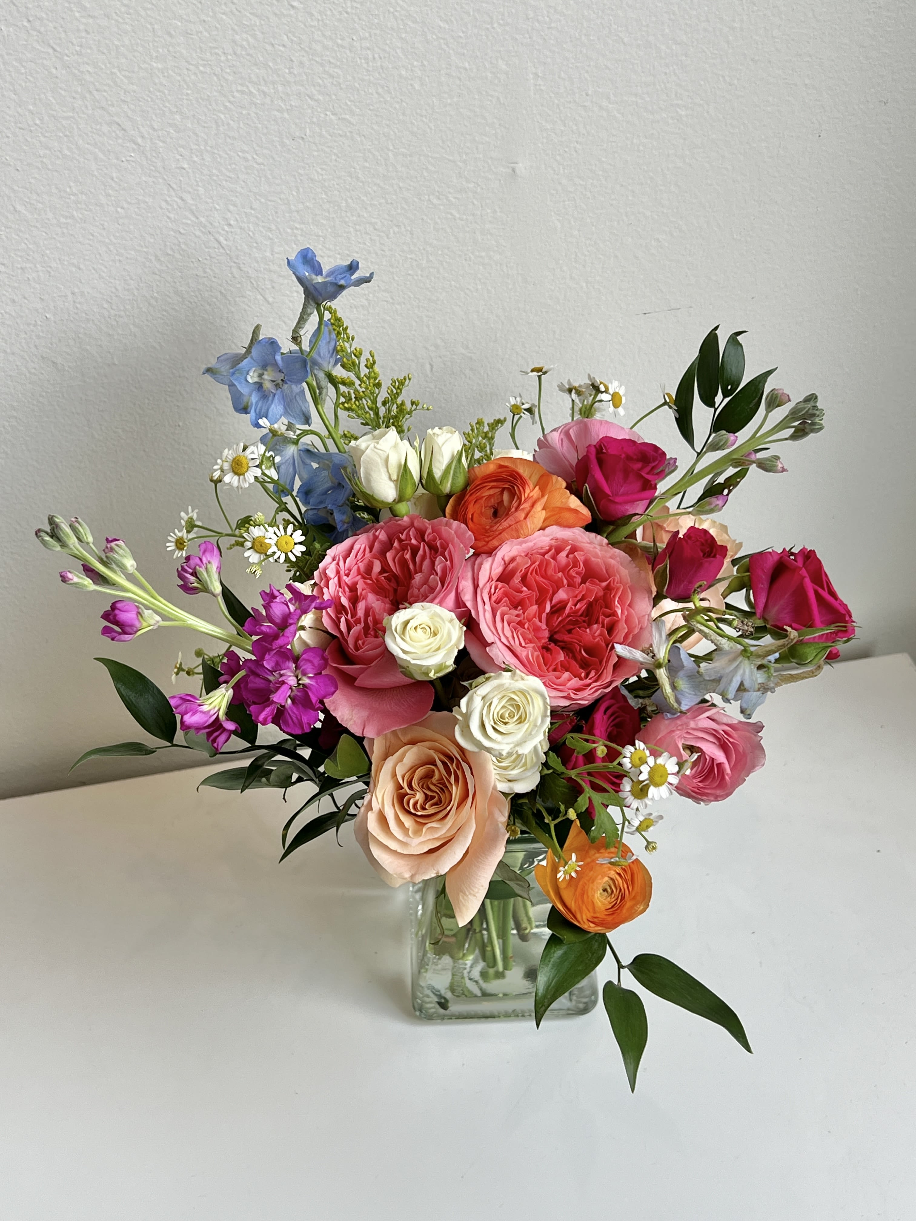 MD26-03 - A Bright Summer Design of Garden Roses, Roses, Delphinium, Ranunculus and Spray Roses With Assorted Foliage and Filler in a Glass Vase.