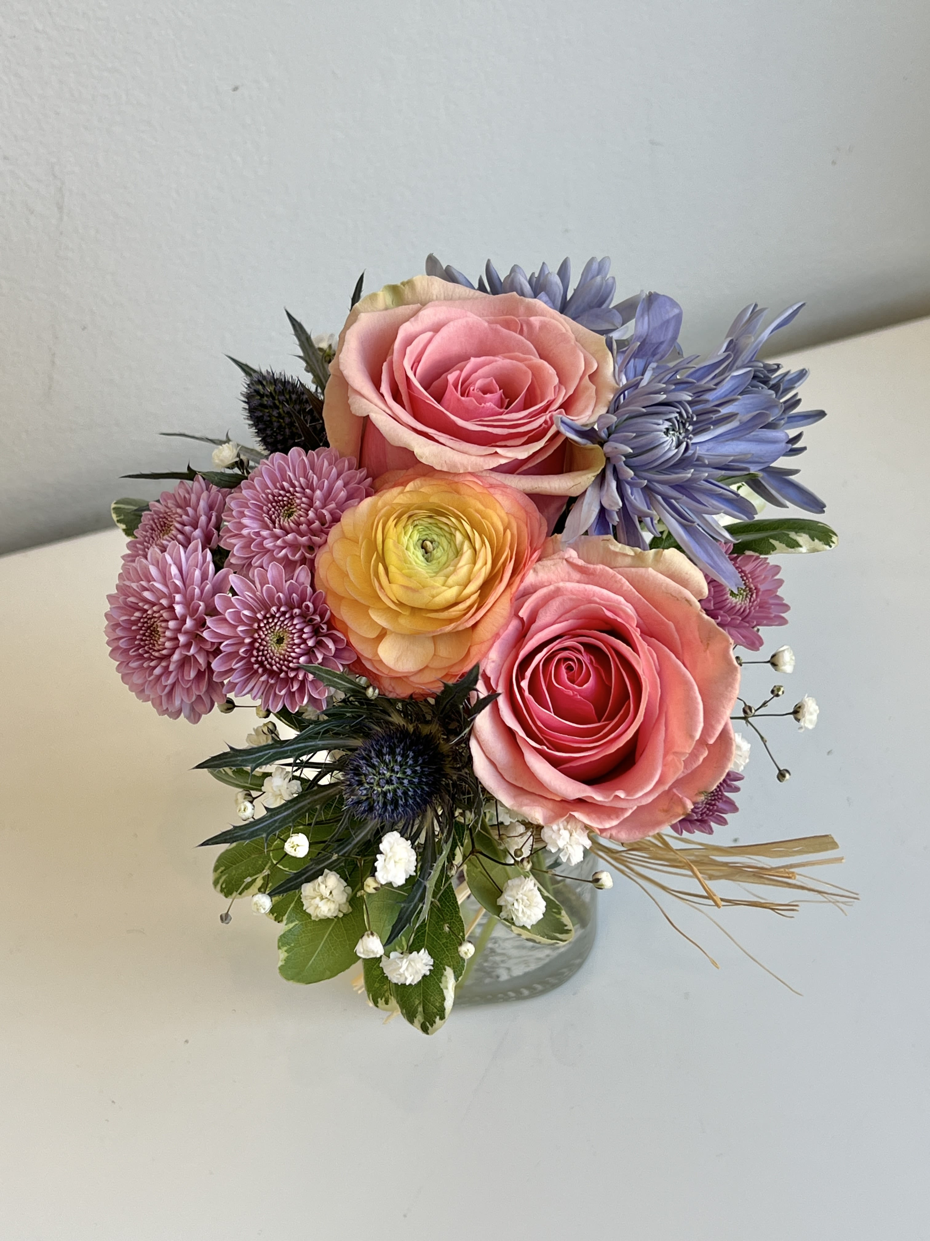 MD26-04 - Pastel Spring Colored Roses, Mums, Ranunculus, Thistle and Baby's Breath With Assorted Foliage in Small Hammered Glass Vase.