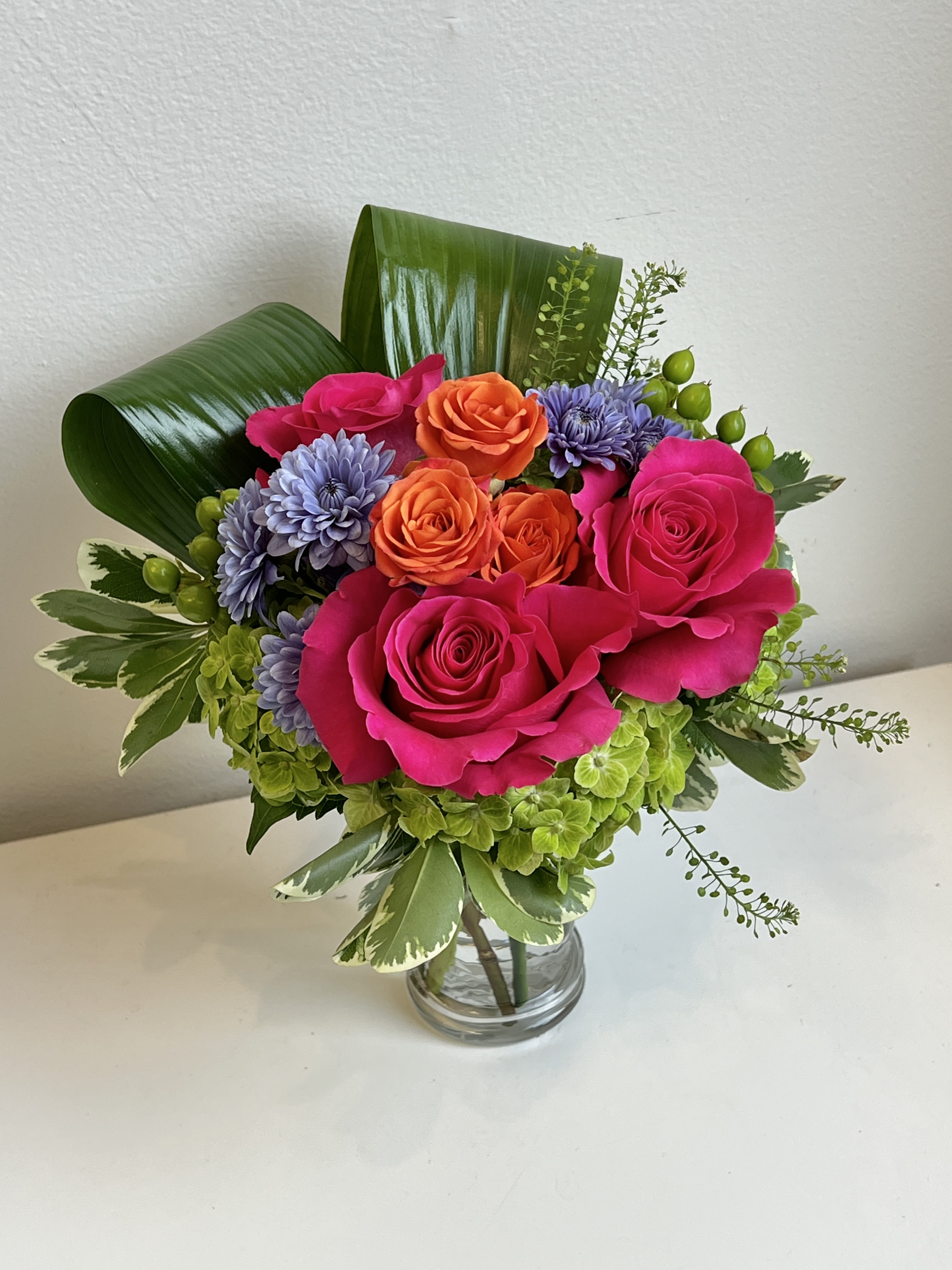 MD26-05 - Bright Bouquet Designed With Green Shamrock Hydrangea, Hot Pink Roses, Orange Spray Roses, Blue Mums, Green Hypericum Berries and Assorted Foliage and Filler in a Glass Vase.