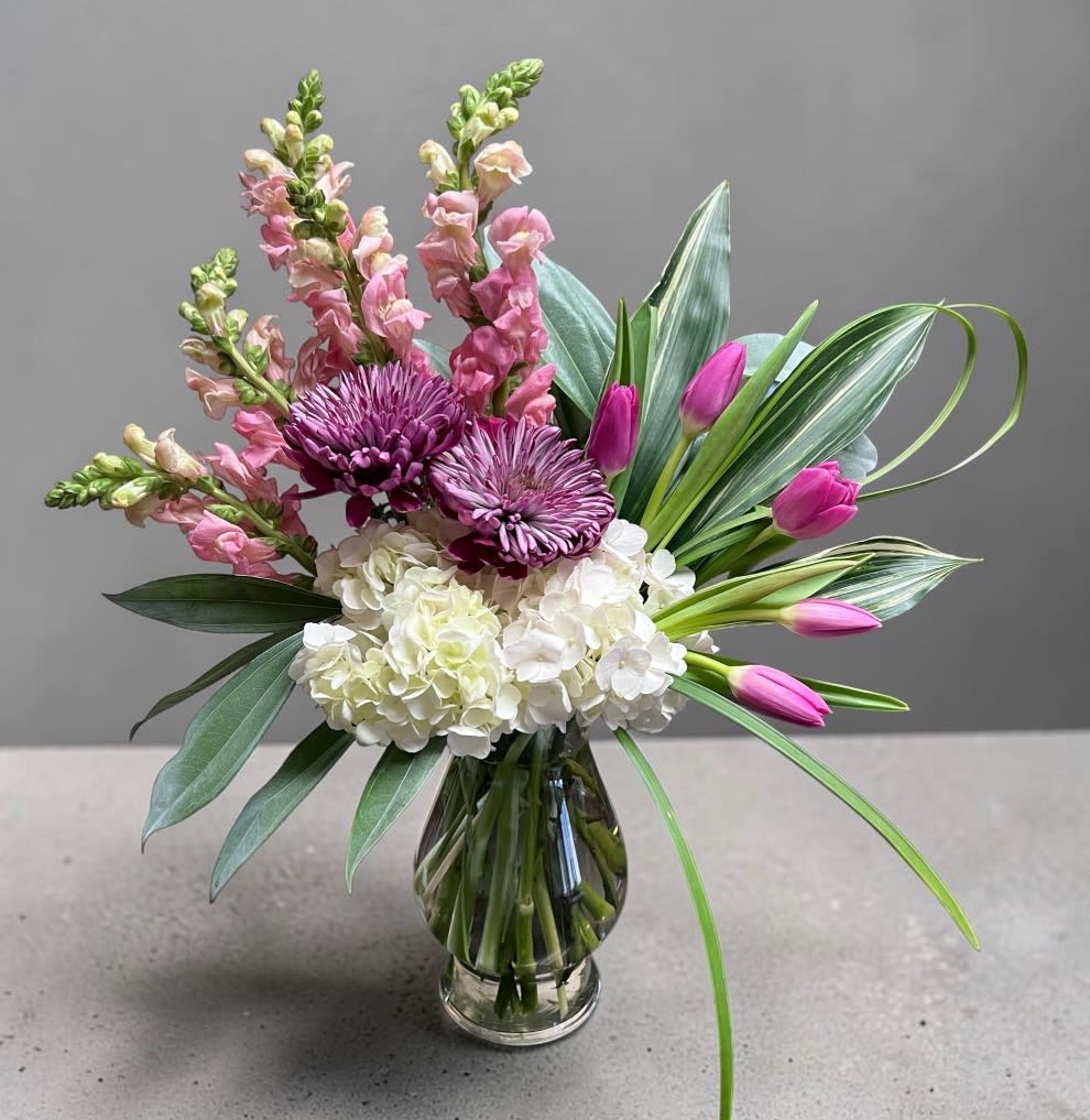 The Layla - The Layla is a soft yet striking design featuring lush white hydrangeas, bold mauve chrysanthemum, delicate pink snap dragons, and vibrant tulips, accented with layered premium greenery for a modern airy finish. Designed in a clear glass vase, this arrangement brings height, texture, and graceful movement to any space.