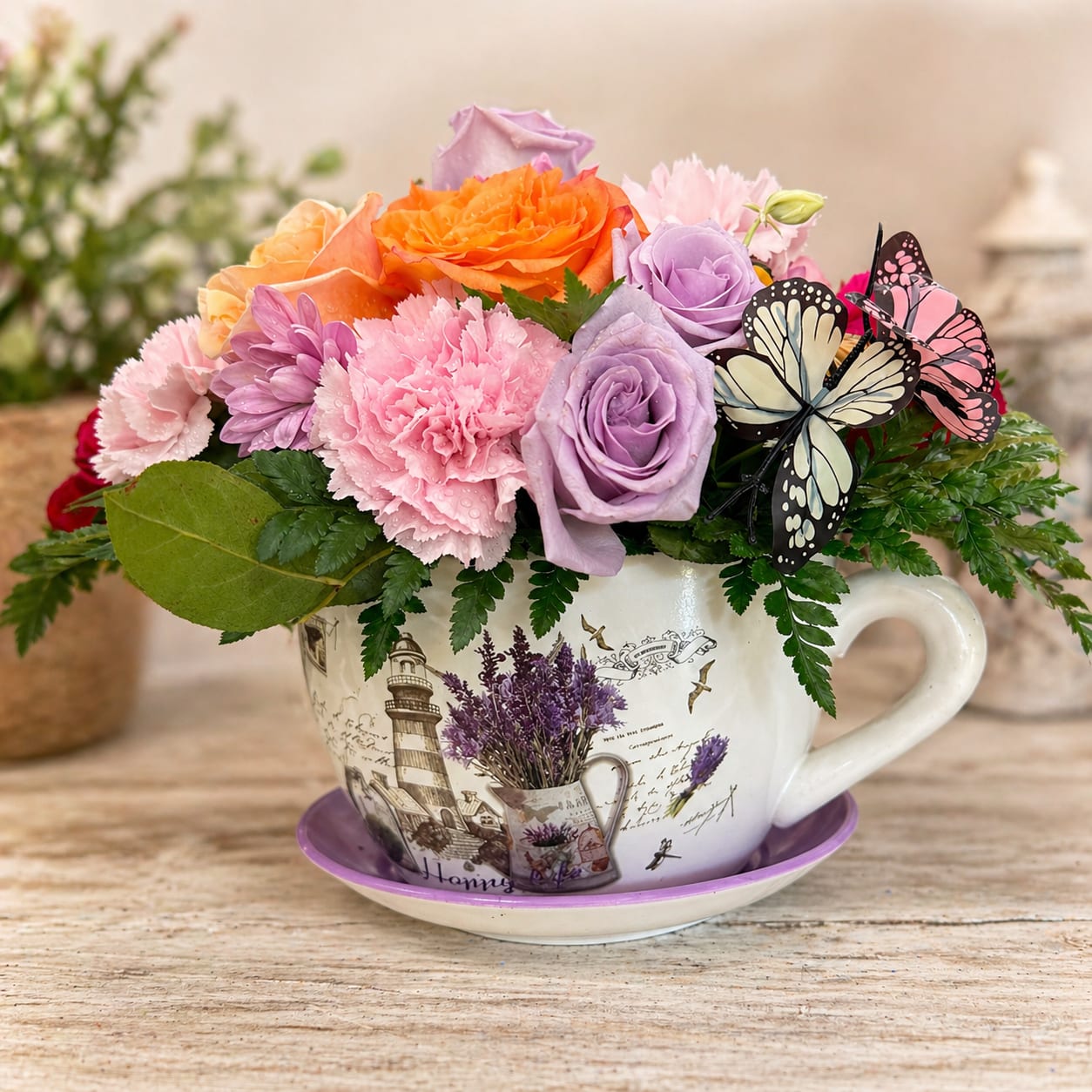 Lavender Dream Cup -  charming and delicate floral arrangement presented in a vintage-style teacup, perfect for adding a soft and elegant touch to any space   This design features a beautiful mix of pastel roses, carnations, and seasonal flowers, complemented with fresh greenery and decorative butterflies for a unique and graceful finish.