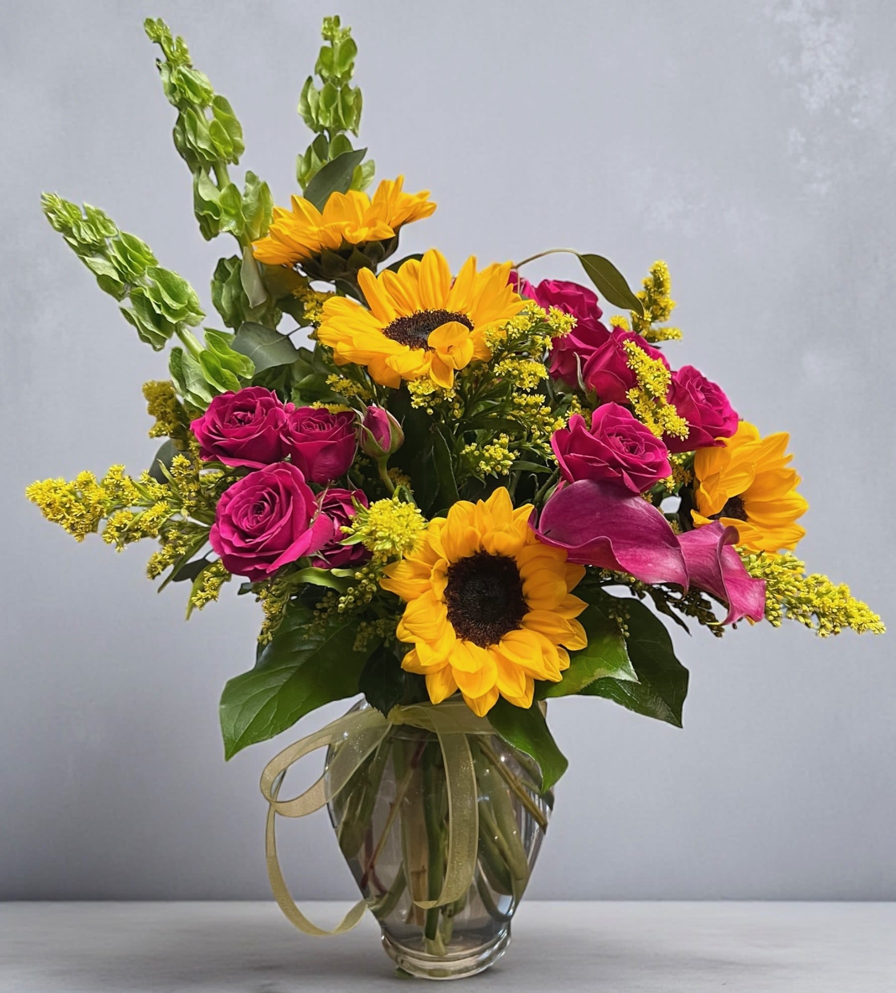 Sun-Kissed Diva - Bright, Bold and Beautifully unforgettable. Sun-kiss Diva is a radiant arrangement bursting with warmth and confidence. Featuring vibrant sunflowers rich fuchsia roses and and golden accent blooms. This design blends sunshine and sass into one stunning bouquet. 