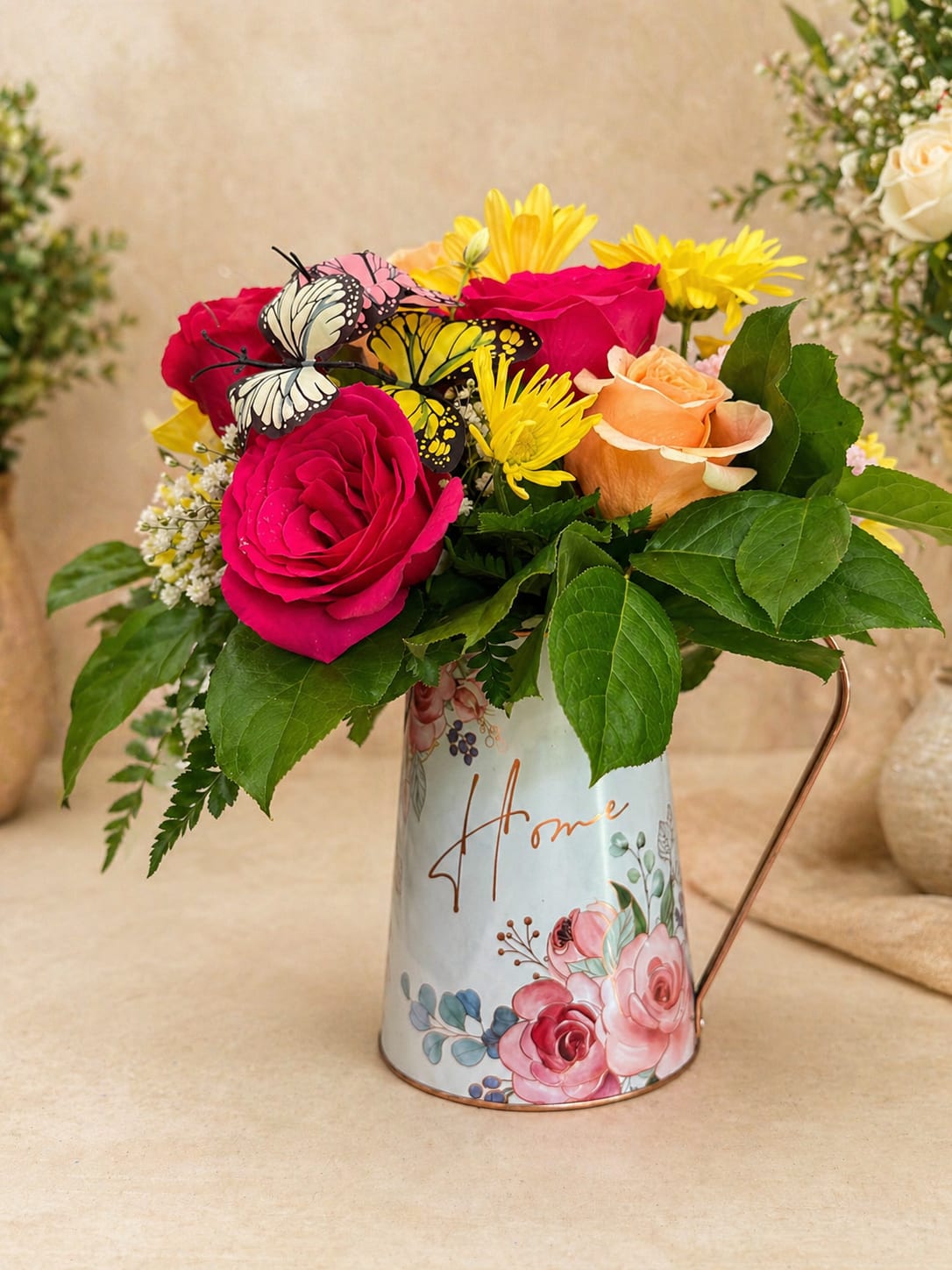 Floral Home Elegance - A warm and charming floral arrangement designed to bring beauty and joy into any home   Presented in a decorative vintage-style pitcher, this arrangement features a vibrant mix of fresh roses, daisies, and seasonal flowers, enhanced with lush greenery and delicate butterfly accents for a unique and elegant touch.