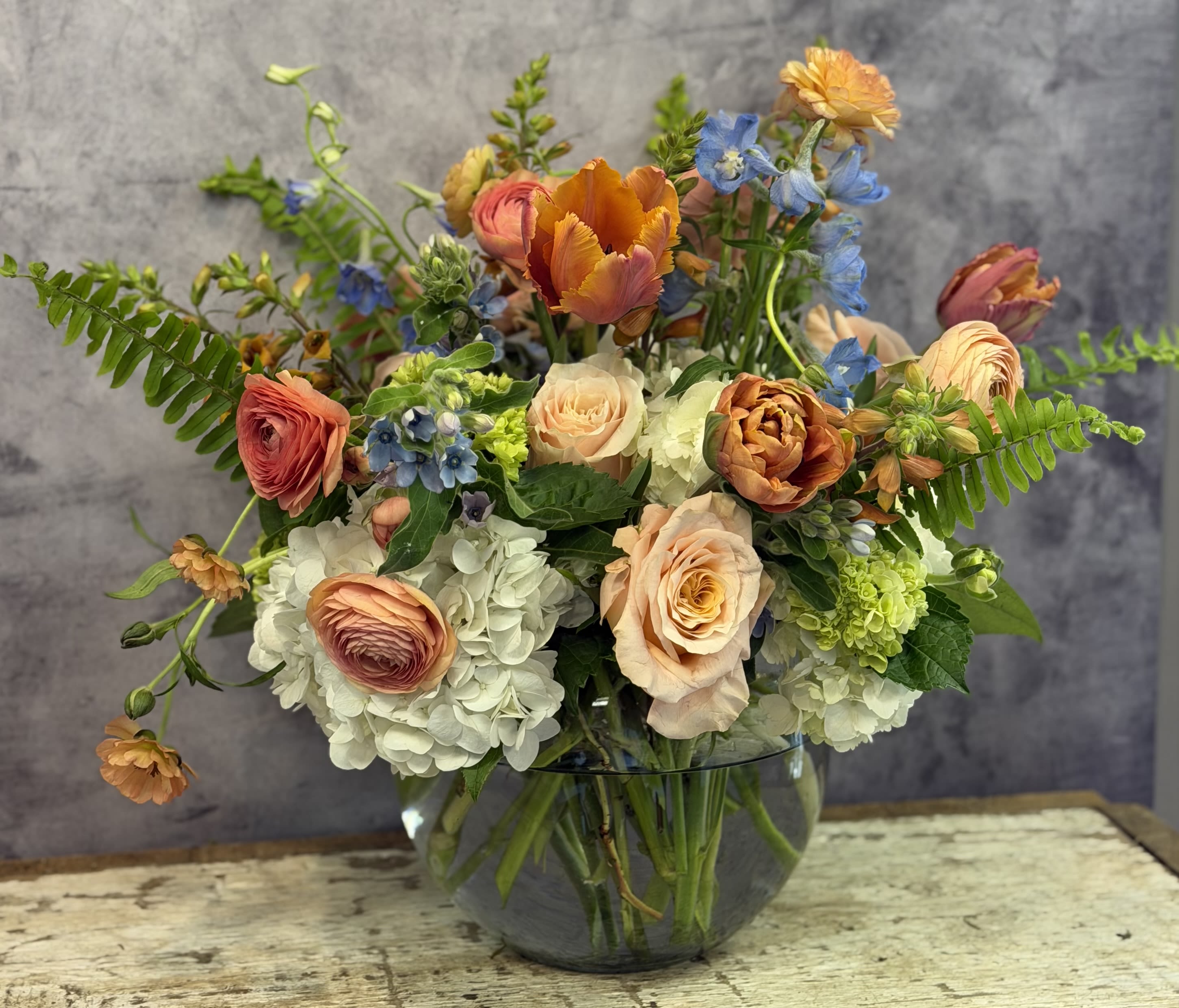 Foxy Mama - Peach and orange roses paired with soft blush ranunculus and a sprinkling of foxglove create the lively, garden-style presence of this mixed-color arrangement. Cool blue delphinium and white hydrangea balance the warm tones, giving the recipient a cheerful, slightly romantic impression the moment they see it.  Arranged in a rounded glass vase with stems visible through the water, the design reads full and layered, spreading broadly across the top with blooms at varying heights for a loose, natural silhouette. Alongside roses, ranunculus, hydrangeas, delphinium, and tulips, the foxglove adds vertical interest and a cottage-garden texture; colors include orange, white, blue, pink, and green for a vibrant, mixed-color palette.  This is the kind of bouquet that makes someone feel genuinely celebrated-bright but thoughtful, casual but elegantly arranged. Delivered in the included glass vase, it arrives ready to display; change the water every few days, trim stems at an angle if needed, and it will keep bringing that fresh, garden-fresh feeling into their home.