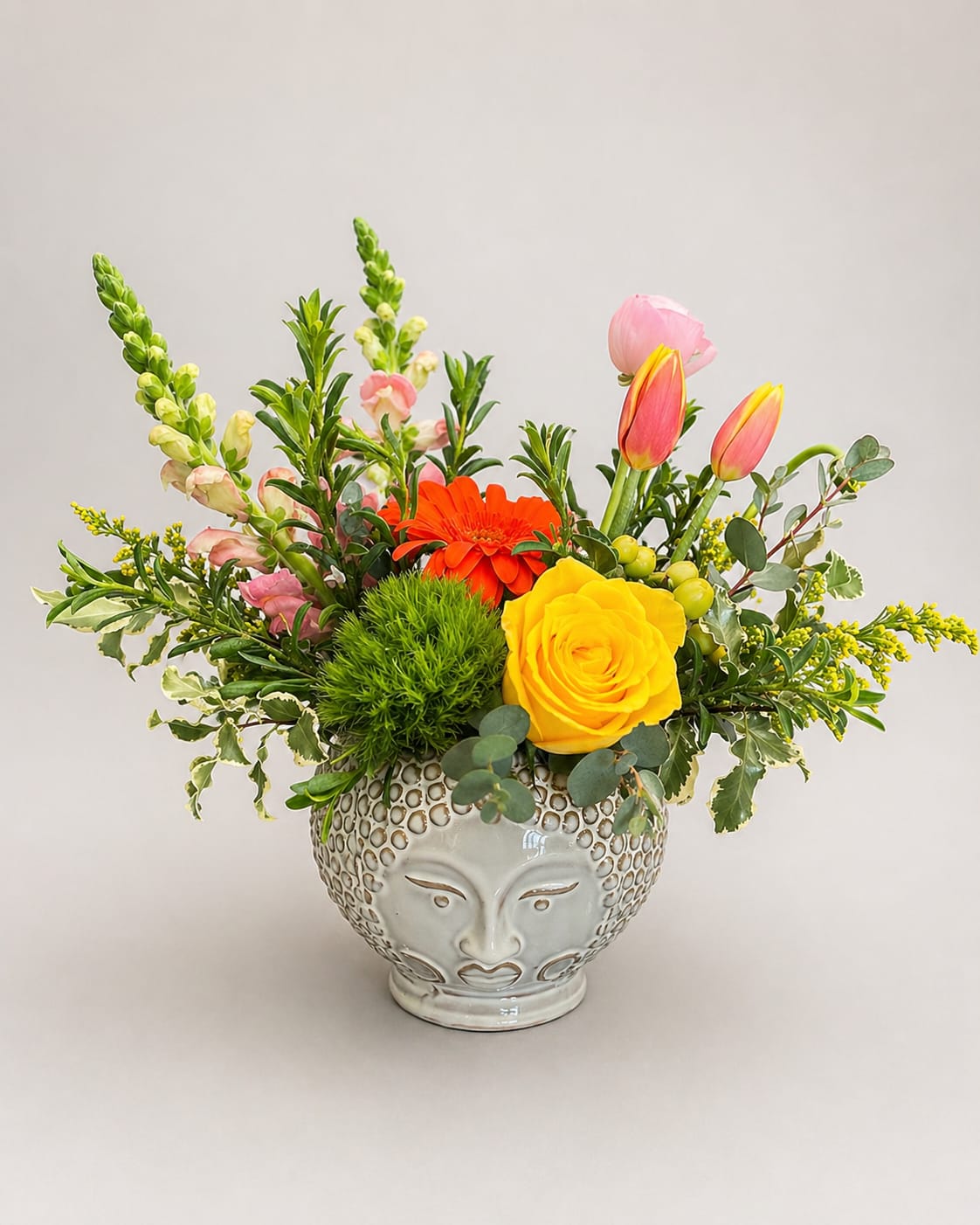 Meditation Garden  - A bright yellow rose and an orange gerbera daisy take center stage in this lively flower arrangement-perfect as a cheerful birthday bouquet, everyday gift, or thank-you arrangement that makes someone feel genuinely celebrated. The product type is a flower arrangement suited for sending warmth and joy; its vivid mixed-color palette of yellow, pink, and orange reads sunny and upbeat, ideal for bringing energy to a kitchen table, office desk, or bedside.  This Meditation Garden arrangement features pink tulips, tall snapdragon spikes, and additional seasonal blooms and rounded green foliage that soften the composition and add texture. Flowers are arranged low and spreading for a relaxed, natural look, and they sit in a decorative ceramic vase shaped with a stylized face for a contemporary, artistic touch. The combination of bold blooms and fresh greenery creates visual contrast and a pleasantly fragrant presence.  Give this to the person you're thinking of when you want them to smile-whether celebrating a birthday, saying thanks, or simply brightening an ordinary day. The low ceramic vase makes it easy to place on a table or shelf; order deadlines and care instructions are provided at checkout so the recipient enjoys peak freshness upon delivery.