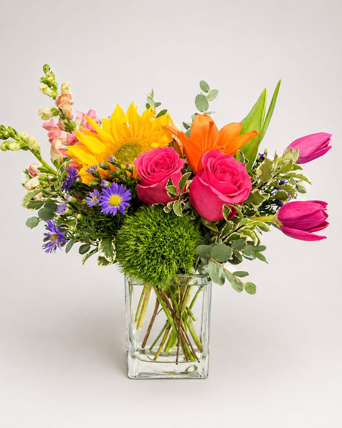 Sunlit Sorbet - A burst of sunlit color-pink roses, a bold yellow sunflower, orange lilies and magenta accents come together in a lively summer bouquet perfect for a birthday, Mother's Day, or "just because" surprise. This flower arrangement reads cheerful and reassuring, making it a thoughtful choice for Get Well wishes, a Housewarming gift, or to say "I'm sorry" or "thank you" in a memorable, uplifting way.  Carefully gathered into a clear glass vase, the design mixes roses, lilies, sunflowers, gerbera-style blooms, snapdragons and small purple aster daisies for a full, rounded garden-style look. The palette of pink, purple, yellow and orange creates vibrant contrast and texture-tall snapdragons add vertical movement while the sunflower anchors the composition. Sized to sit beautifully on a dining table or bedside, the arrangement arrives ready to display; refresh the water and trim stems every few days to keep flowers lively for longer.