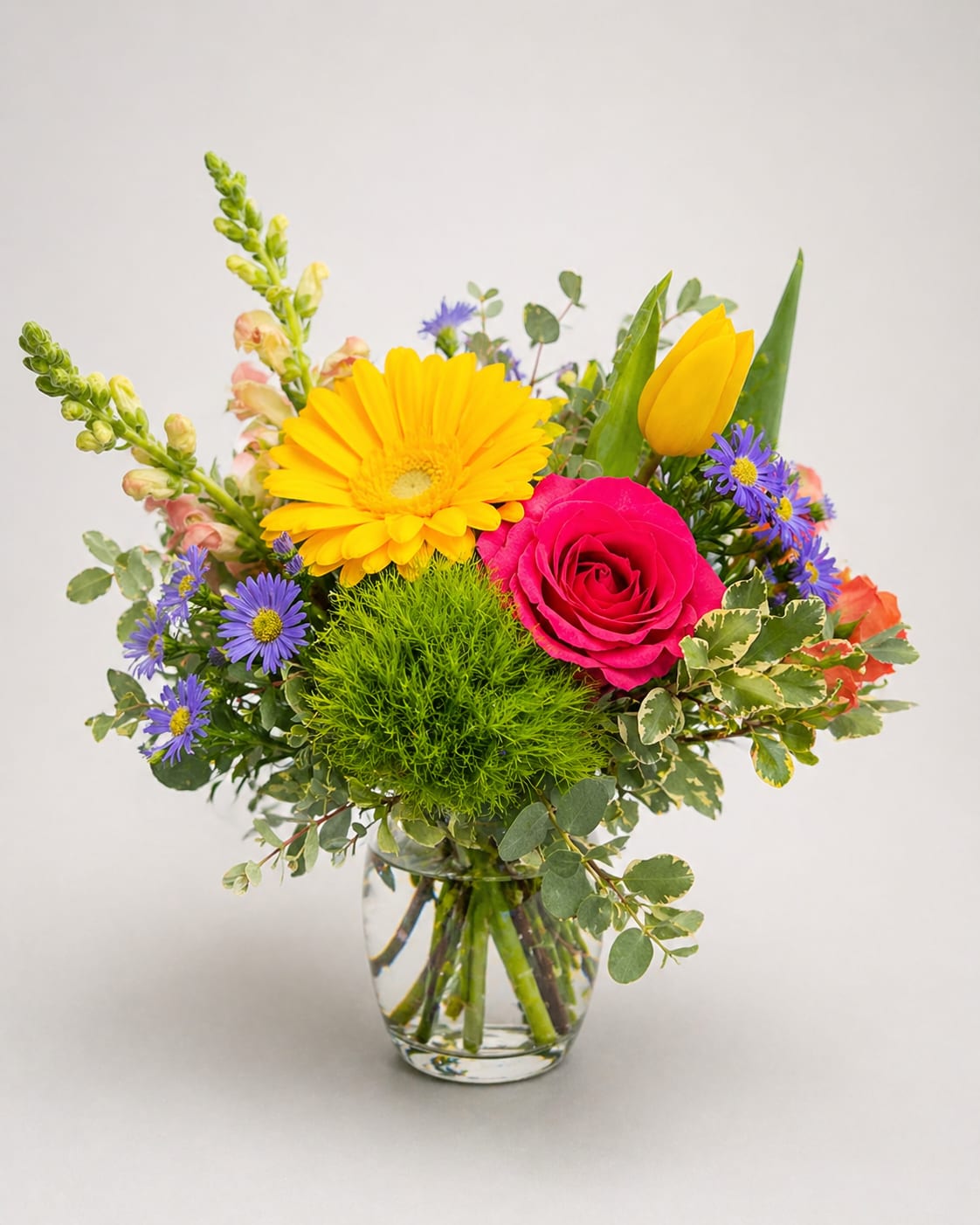 Summer Splash  - A bright yellow gerbera daisy and a vivid pink rose take center stage in this Summer Splash flower arrangement, making it an ideal birthday bouquet, everyday gift, or cheerful thank-you arrangement. The mix of warm focal blooms and cool purple accents gives the recipient an immediate lift-she'll feel noticed and celebrated the moment the vase arrives.  This loose, airy design pairs yellow tulips, tall snapdragon spikes, and daisy-like purple asters with soft pink roses and gerbera blooms for lively contrast and texture. Arranged in a clear glass vase with visible stems, the casual presentation reads fresh and unfussy while still feeling thoughtfully styled; colors include mixed tones of yellow, pink, and purple.  Perfect for brightening a kitchen table, office desk, or bedside, Summer Splash suits anyone who loves playful color and garden-fresh energy. Vase included; to keep blooms lively, trim stems and refresh water every 2-3 days and place away from direct heat.