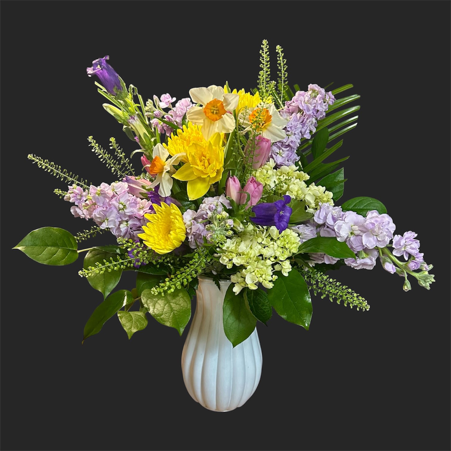 All Things Bright  - A sunny, spring-forward bouquet that feels like a garden in full bloom-this flower arrangement is crafted as a cheerful birthday bouquet or seasonal gift to brighten someone's day. The mix of yellow daffodils and chrysanthemums leads with joyful color, while lavender stock and pink tulips add a soft, romantic contrast that makes the recipient feel truly celebrated.  Arranged in a white ribbed ceramic vase, the design reads airy and abundant: broad glossy foliage fans outward for a full silhouette, and delicate white accent blooms give the whole composition a refined finish. Colors include yellow, purple, white, and pink; flowers include daffodils, chrysanthemums, stock, tulips, and complementary seasonal stems for texture.