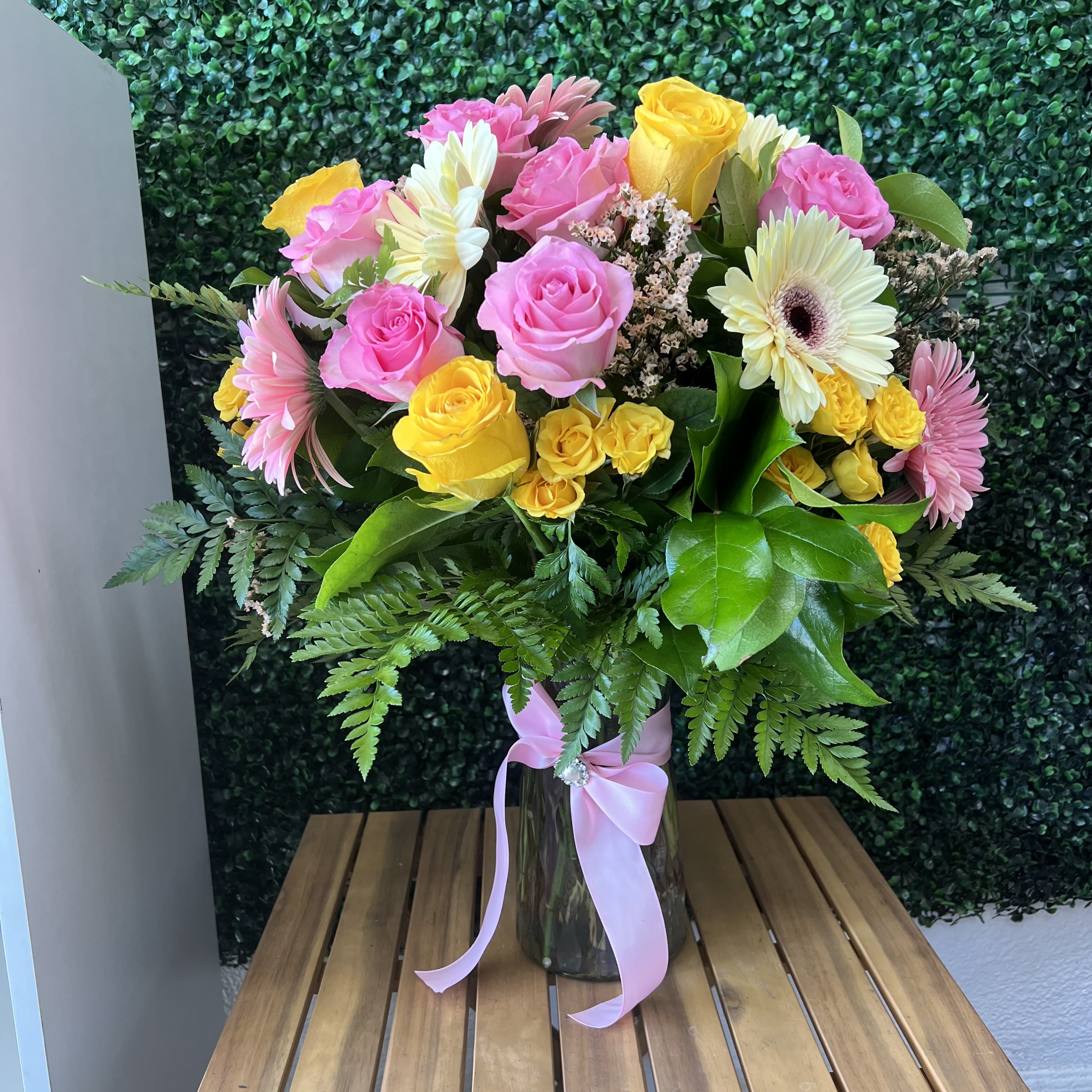 Sunshine Blush Garden Vase - A cheerful and elegant mixed arrangement featuring radiant yellow roses, soft pink roses, blush gerbera daisies, creamy white blooms, delicate filler flowers, and lush greenery, beautifully designed in a clear glass vase finished with a pink satin ribbon. This vibrant bouquet brings warmth, joy, and sweet charm—perfect for birthdays, celebrations, thank you gifts, or simply brightening someone’s day.