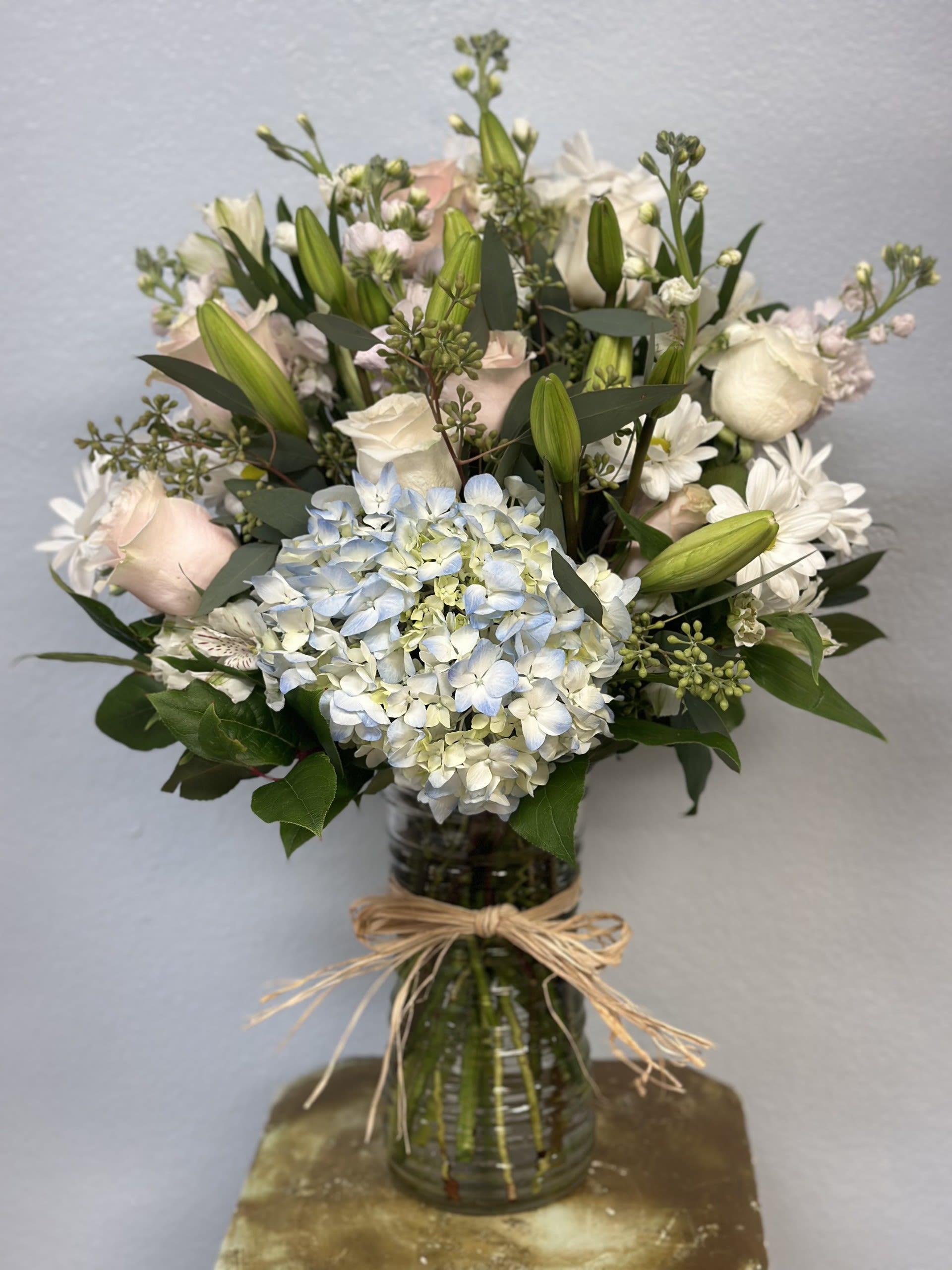 Soft Serenity Blooms - A pale blue hydrangea anchors this Soft Serenity Blooms arrangement, surrounded by white roses, lilies, alstroemeria, and daisy-like blooms for a calming birthday bouquet, sympathy arrangement, or wedding centerpiece. Cool blue, white, and blush accents set against deep green foliage create an elegant, serene mood that comforts the person you're thinking of when words feel small.  The full, rounded design has upright lily buds for graceful height and a slightly formal silhouette. Arranged in a clear glass vase and finished with a raffia bow, the mix of cloudlike hydrangea clusters, smooth rose petals, and crisp alstroemeria feels airy and refined. Vase-ready and easy to display-keep in fresh water, away from direct heat, and recut stems to extend vase life.