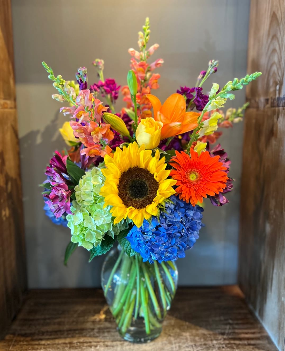 Bright mixed bouquet with sunflower, gerbera daisy, hydrangea, and snapdragons in a glass vase